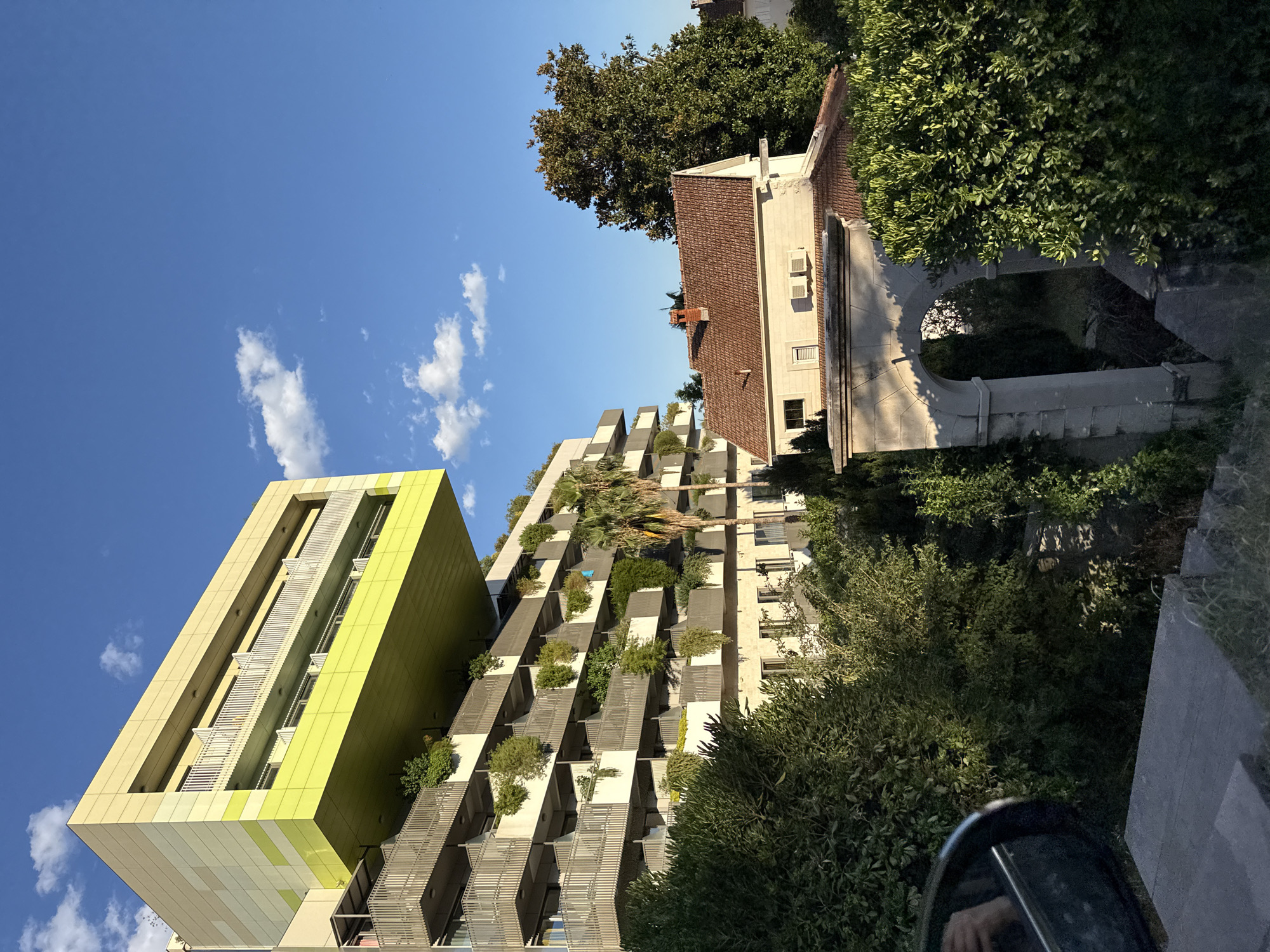 The image shows a modern apartment building with a striking, cantilevered section clad in yellow and lime green panels. The building has numerous balconies adorned with planters overflowing with greenery. To the right of the modern structure, a traditional-style house with a terracotta tiled roof is visible, featuring an arched stone entryway in the foreground. The scene is bathed in the warm light of late afternoon or early evening, casting shadows and highlighting the textures of the buildings and foliage. Lush trees and shrubs surround the properties, creating a sense of integration with nature. The sky is a clear, vibrant blue with a few scattered white clouds. The location is Jošice, Montenegro.