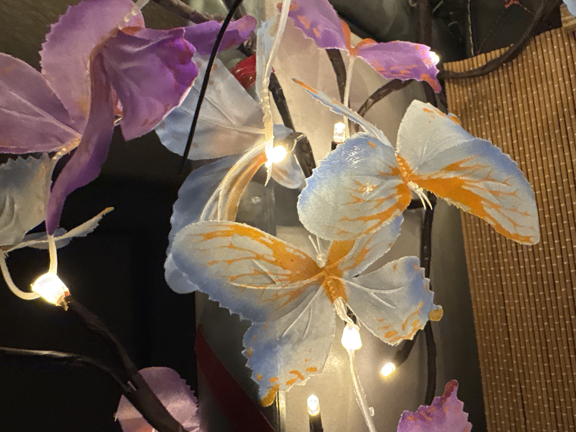 The image shows a close-up of decorative artificial butterflies with blue and orange wings, interspersed with purple floral decorations and illuminated by small, warm white LED lights. The butterflies and decorations are attached to dark, thin branches. In the background, a dark, reflective surface is visible, possibly glass or a dark painted wall, with glimpses of red and white objects. The foreground features a textured, woven brown surface, resembling a mat or a placemat. The scene is set indoors, with no specific indicators of a location in Osterode am Harz, Germany. There are no people or animals present in the image.