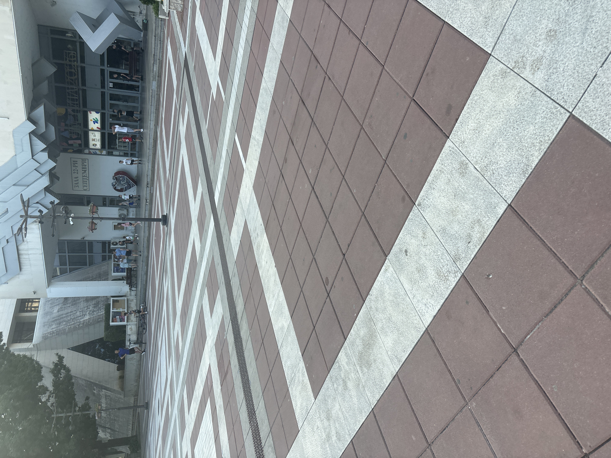 This detailed image captures an outdoor public plaza in Blagoevgrad, Bulgaria, specifically in front of the "Zala 22-ri Septemvri" (Hall 22nd September) and the "Naroden Teatar" (National Theater).
The foreground and midground feature a paved surface composed of square tiles arranged in a repeating geometric pattern of dark reddish-brown and light gray. A metallic drainage grate runs horizontally through the mid-section of the plaza. A multi-armed black lamppost with hanging plant baskets stands on the left side of the midground.
In the midground, several individuals are present. On the left, a man wearing a blue shirt and dark shorts is walking or jogging, while a boy in a black and white striped shirt stands near a bicycle that is lying on its side on the pavement.
The background is dominated by two distinct architectural structures. To the right, the main building, identified by the Cyrillic text "ЗАЛА 22-РИ СЕПТЕМВРИ" (HALL 22ND SEPTEMBER) and "НАРОДЕН ТЕАТЪР" (NATIONAL THEATER) on its facade, exhibits a modern design with a complex, angular roofline and a large glass entrance area. A large, gray, abstract, L-shaped sculpture stands to the right of the entrance, and a red heart-shaped art installation is visible to the left of the main doors. Numerous people are gathered near this building's entrance, standing, walking, or resting on dark benches. One person in a white t-shirt appears to be stretching or adjusting clothing.
To the left of the main building is another structure with a distinctive, starker architectural style, featuring a prominent triangular cutout on its side. Trees with green foliage are visible along the far left, partially obscuring this building and casting some shade. The overall scene depicts a lively public square on a clear day, with people engaging in casual activities.