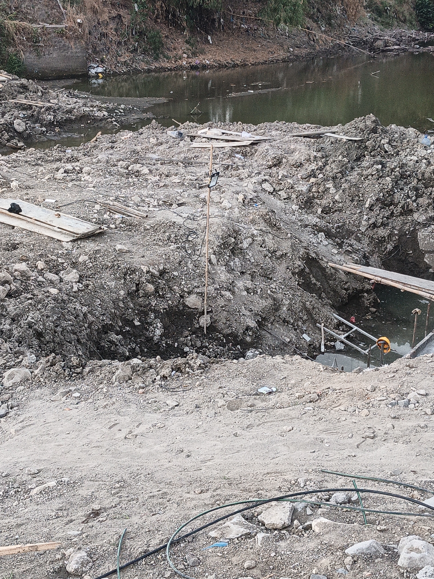 The image depicts a construction site near a body of murky water, likely a river or canal, with lush vegetation and debris along its banks. The foreground is dominated by piles of excavated soil and rocks, indicative of ongoing earthmoving activities. Several wooden planks and metal pipes are scattered across the site, suggesting the presence of construction materials. A dark, irregularly shaped hole in the ground, filled with water and mud, is prominent in the middle ground, possibly a result of excavation or drainage. Construction equipment, including what appears to be a safety helmet, is visible near the water's edge. The overall scene suggests a scene of urban development or renovation in a potentially underdeveloped area, with the water body serving as a natural boundary. The location is identified as Planggu, Indonesia, which suggests a tropical or sub-tropical climate, consistent with the dense vegetation.