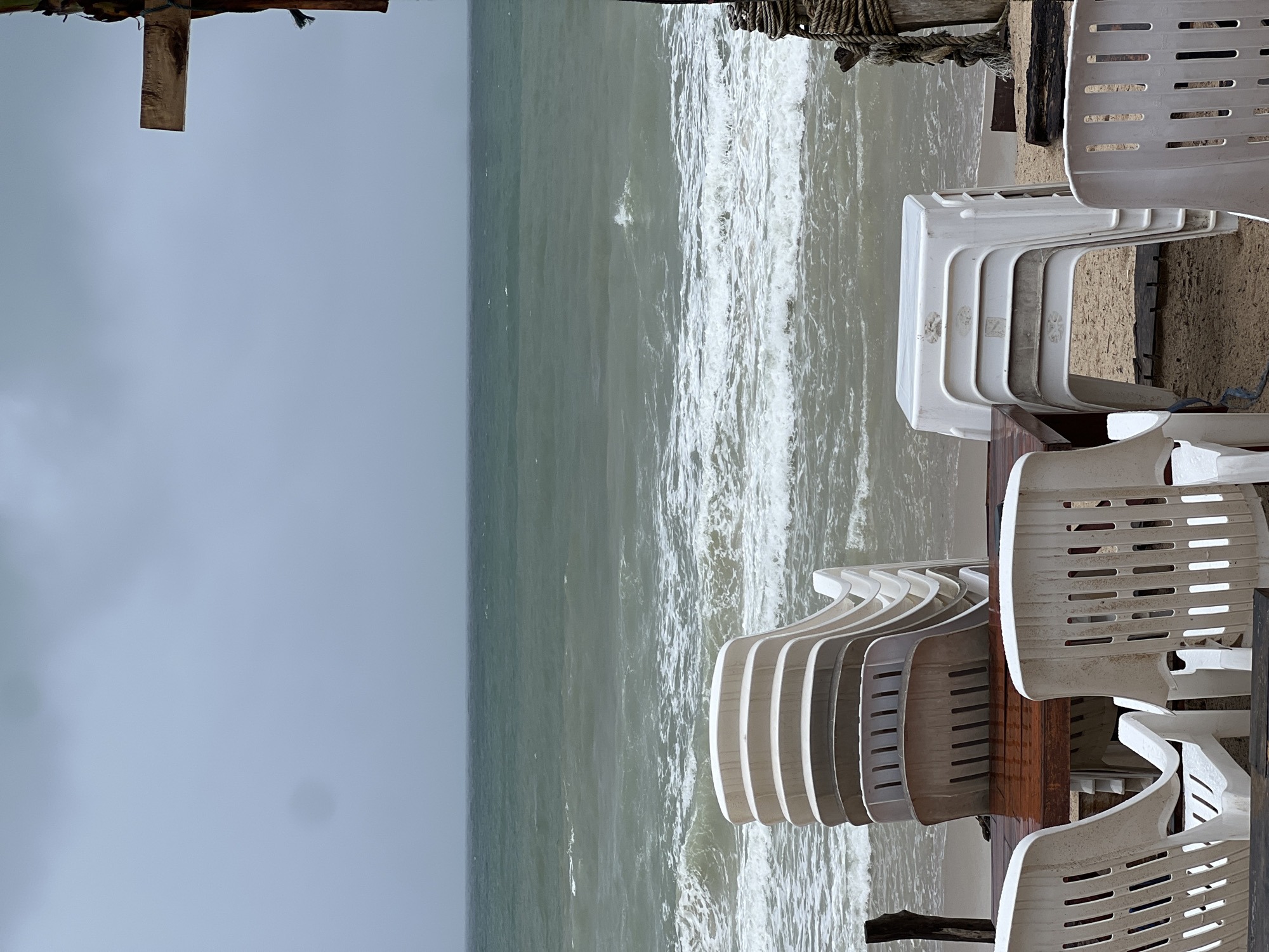 The image depicts a quiet beach scene on an overcast day, viewed from a slightly elevated perspective. The primary subjects are the ocean and numerous white plastic chairs positioned along the shore or on a sandy platform.
In the foreground and to the right, several stacks of white plastic chairs are neatly arranged. Two main stacks are prominent: one stack of five or more chairs faces towards the left, and another stack of similar chairs is positioned further back. Additional individual chairs, some partially visible and others facing away, are scattered across the right side of the frame. These chairs appear to be made of white, somewhat weathered plastic, with slatted backs and seats. Faint, illegible markings or dirt are visible on some chair surfaces.
The mid-ground is dominated by the ocean, which features a greenish-grey hue under the subdued light. Numerous white-capped waves are visible, actively breaking as they roll towards the unseen shore. The sea surface appears choppy.
The upper-left portion of the image shows a uniformly light grey, overcast sky, indicating cloudy weather or a lack of direct sunlight. A wooden beam or plank, possibly part of a pier, overhead structure, or roof, extends into the frame from the top edge. Thick, coiled ropes are visible around other wooden elements in the top-right, suggesting mooring or structural supports.
No people are present in the scene, contributing to a sense of tranquility or suggesting the location is not currently in use, possibly off-season or before/after operating hours. The weather appears cool and muted due to the heavy cloud cover. There is no clearly visible, legible text in the image.