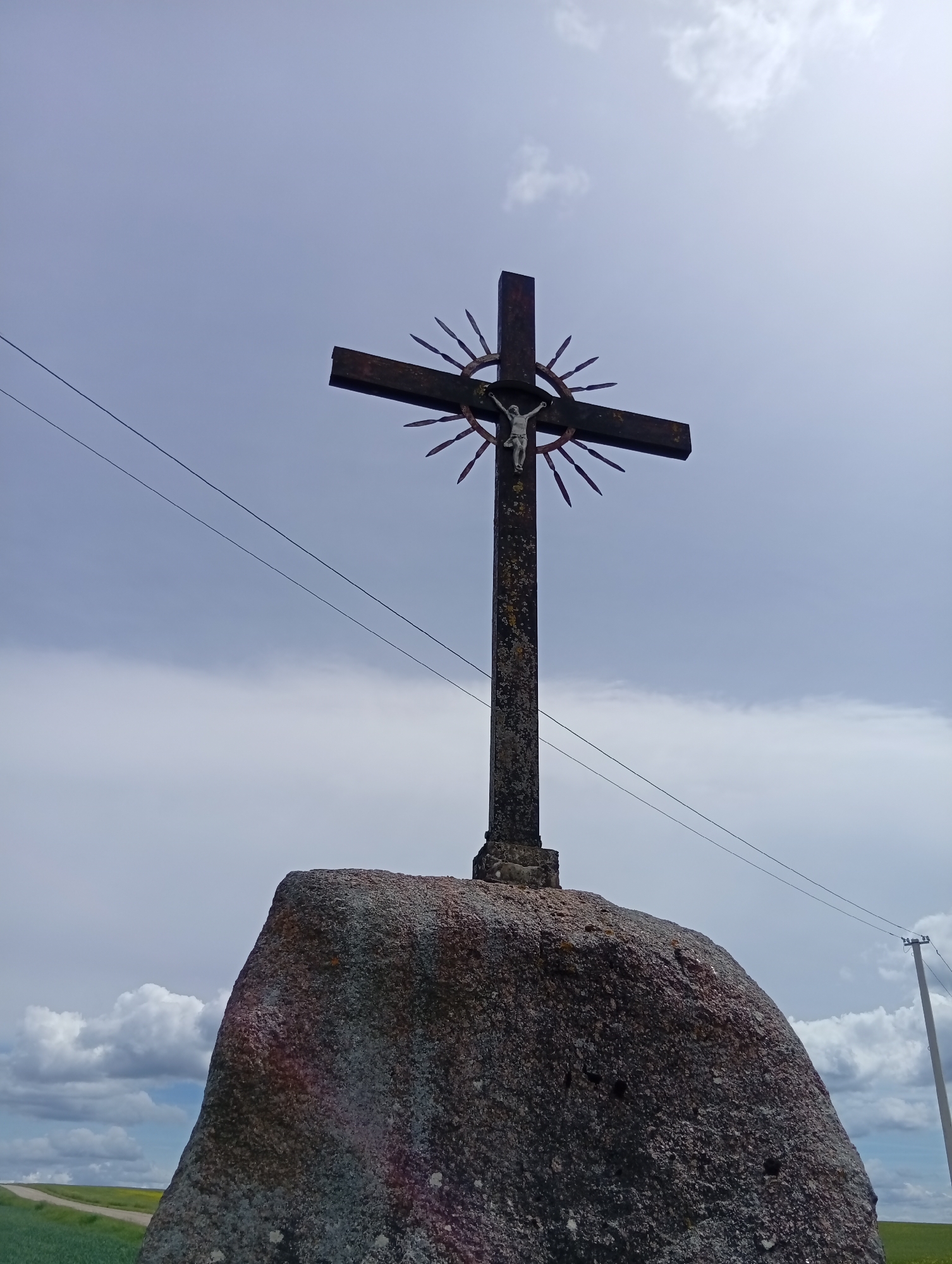 A tall, weathered stone cross is erected on top of a large boulder. The cross itself is dark and appears to be made of metal, with a figure of Jesus Christ affixed to it. A circular metal halo with spikes emanates from the cross. The scene is set against a cloudy sky. In the background, power lines stretch across the frame, and a utility pole is visible to the right. To the left, a dirt road or path leads through a green field. The location is identified as Krikliai, Lithuania.