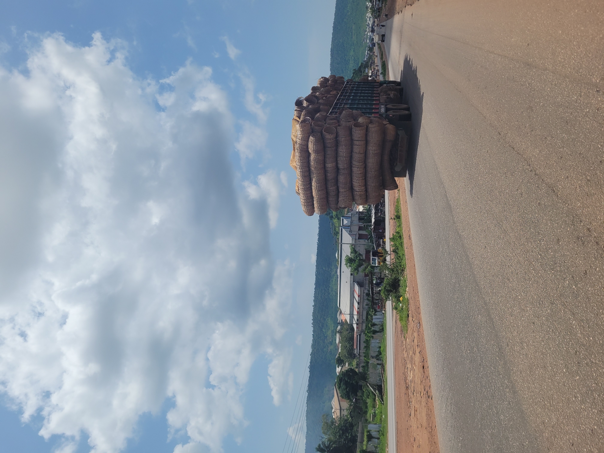 A large truck loaded with cylindrical, straw-colored bundles is traveling on a paved road in Lokoja, Nigeria. The truck is positioned in the right lane of a multi-lane highway, moving away from the camera. The bundles appear to be tightly packed and secured with netting and a large piece of beige tarp. The truck has a dark green frame and dual rear wheels on both axles. In the background, there are green, tree-covered hills under a bright blue sky with scattered white clouds. Along the side of the road, there are various buildings, including structures with corrugated metal roofs, as well as some vegetation and other vehicles. The road surface is asphalt and shows signs of wear.