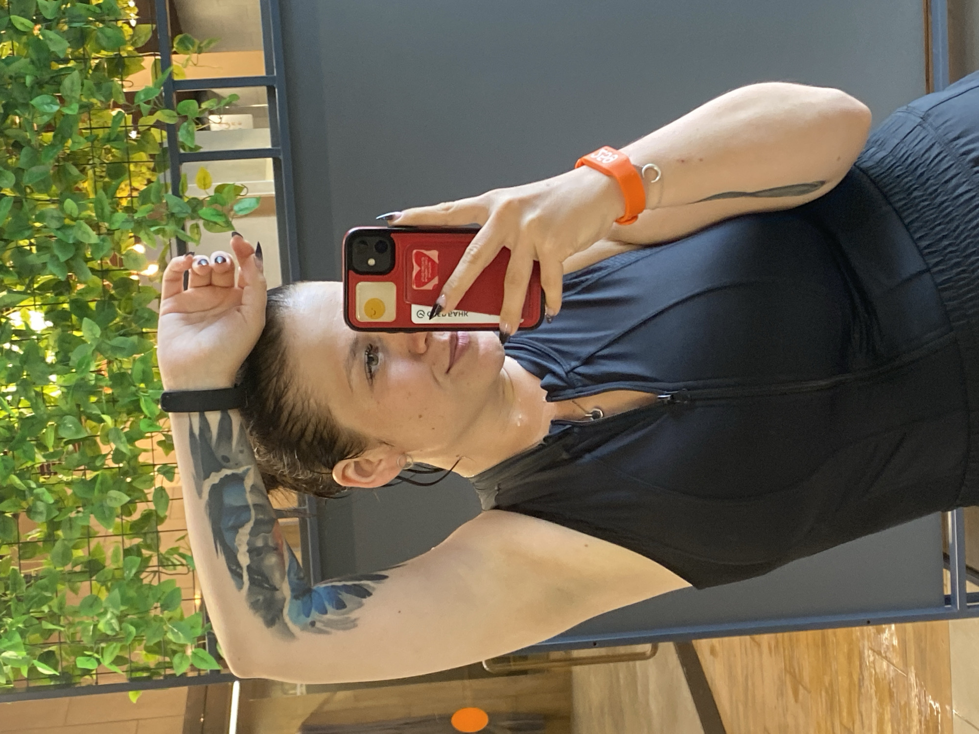 The image shows a woman taking a selfie in what appears to be an indoor gym. She is wearing a black sleeveless workout top with a zippered front and black shorts. She has tattoos on her left arm, including a bird-like design. On her left wrist, she wears a black fitness tracker, and on her right wrist, an orange wristband. She is holding a red smartphone in her right hand, which is pointed towards herself to capture the image. In the background, there is a decorative plant wall with green foliage and string lights, and a grey partition. The flooring visible in the lower left corner appears to be wooden. There is no indication that this scene is located in Votkinsk, Russia.