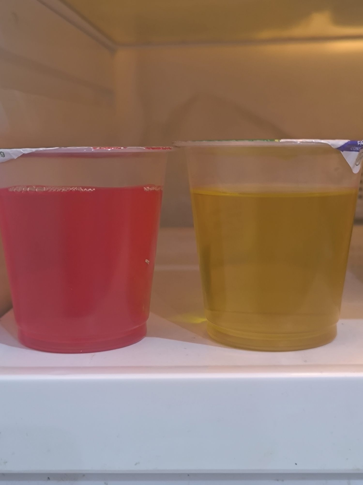 The image shows two clear plastic cups filled with liquid, placed side-by-side on a white surface. The cup on the left contains a vibrant red liquid, while the cup on the right holds a yellowish liquid. Both cups appear to be single-serving containers with some form of foil or plastic lid covering the top, partially peeled back on the right cup. The background is a muted, warm tone, suggesting an indoor environment, possibly a refrigerator or pantry shelf. There are no people, animals, or explicit signs of a specific location within Chișinău, Moldova. The overall scene is simple and domestic, focusing on the two beverages.