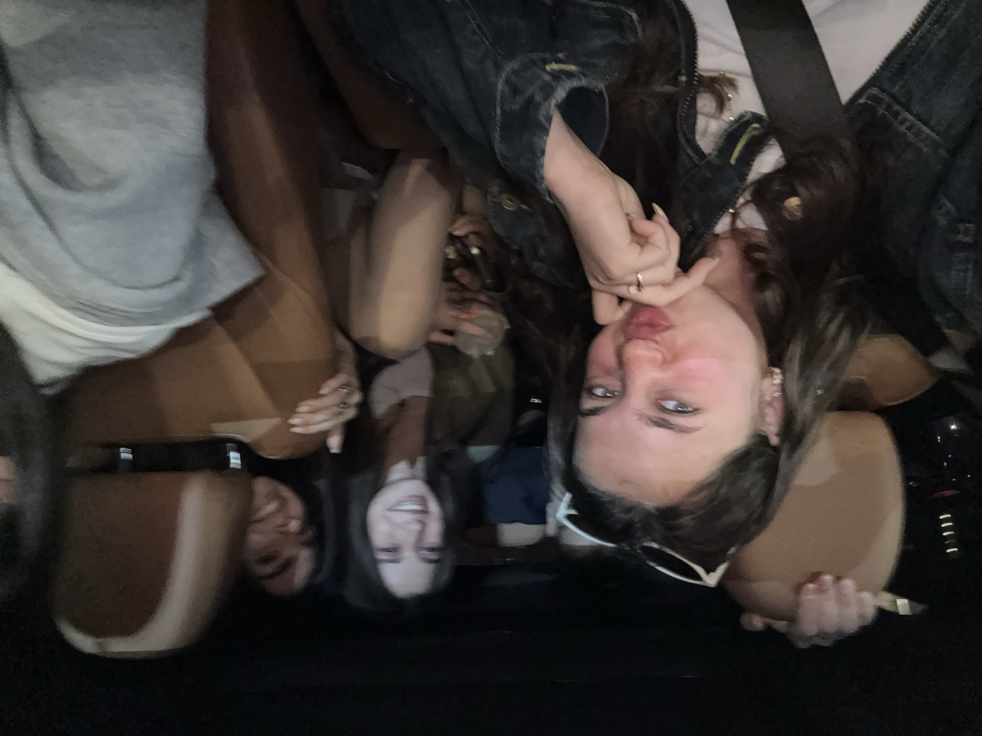 The image captures a casual moment with three young women inside what appears to be a car, likely in Yerevan, Armenia. The photo is taken from an inverted perspective, showing the subjects upside down, suggesting a selfie or an unusual camera angle.
In the foreground, a woman with long dark hair is prominent, wearing a denim jacket over a light-colored top. She gazes towards the camera with a playful, slightly pouting expression, her left hand adorned with rings and long nails, held near her chin. White-framed glasses are pushed up on her head or around her neck. To her immediate right (viewer's left in the inverted image), another woman with dark hair is visible, smiling warmly towards the camera. A third person's head is partially seen further to the left (viewer's right). In the background, to the far left, a fourth individual is partially visible, wearing a light grey t-shirt over a white undershirt.
The setting is clearly a car interior, indicated by the dark and brown/tan upholstered seats and visible seatbelts. The lighting is dim, suggesting it might be nighttime or a low-light indoor environment. The overall mood conveyed is casual and friendly, with the subjects appearing to be enjoying their time together. No discernible text is visible within the image.