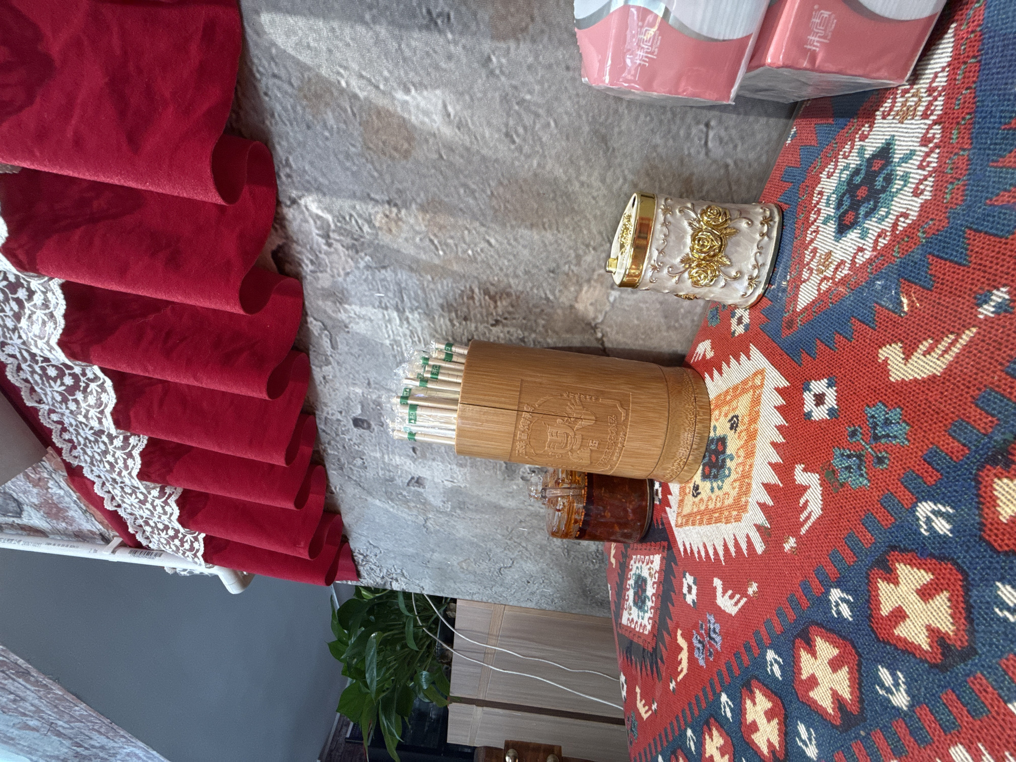 The image depicts a close-up view of a table setting, likely in a restaurant or dining area, in Huizhou, China. The foreground is dominated by a vibrantly patterned tablecloth featuring a red, blue, and yellow geometric design reminiscent of traditional ethnic textiles. Resting on the tablecloth are several objects. To the left, a small glass jar contains a reddish-brown condiment, possibly chili oil or sauce, with a spoon inserted. Next to it, a cylindrical bamboo container holds a bundle of chopsticks, each individually wrapped in clear plastic with green labels. In the center-right, a decorative ceramic dispenser with a gold lid and embossed rose pattern stands. To the far right, two stacks of pink packaged tissues are visible. Behind the table, a textured grey wall serves as the backdrop. Above the wall, a red valance with white lace trim hangs, partially obscuring a white pipe. A green potted plant is visible to the left, behind the table setting. The lighting suggests an indoor environment with some natural light entering from the left. No people are present in the frame.