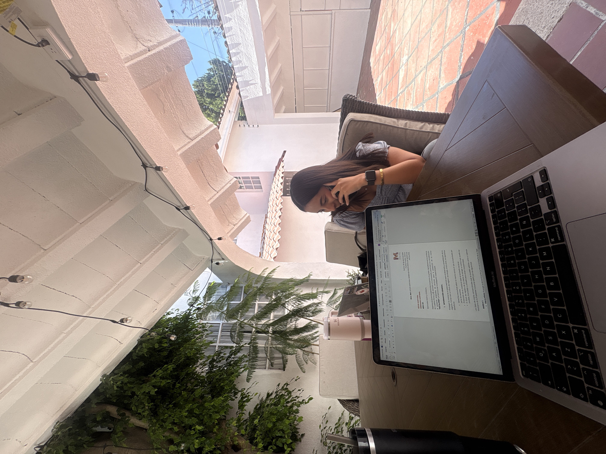 The image depicts a woman seated at a wooden table on an outdoor patio, engaged in a phone call while a laptop is open in front of her. She has long, dark hair and is wearing a light-colored, possibly patterned, top. An Apple Watch and a bracelet are visible on her left wrist as she holds the smartphone to her right ear, appearing focused.
The setting is a sunlit outdoor space, likely a courtyard or patio, with white stucco walls, arches, and reddish-brown tiled flooring visible in the background. Green foliage, including trees with fine leaves and dense bushes, surrounds the area. String lights are strung along the overhead arches, suggesting the patio is also used in the evenings. The bright light and clear shadows indicate a sunny daytime environment.
On the table, a silver MacBook Air laptop is open, displaying a document. A dark-colored tumbler or water bottle is also present near the laptop.
Text visible on the laptop screen includes the title "PROPUESTA DE SERVICIOS" (Service Proposal) for "METALPRO." Other discernible text sections are "KEVIN SANTANA," "RIDIANA SANTA," "MEX-SD," and a date "Jueves, 30 de abril de 2005" (Thursday, April 30, 2005). The document's content discusses "INFORMACIÓN GENERAL" (General Information), "Derechos exclusivos sobre la misma contra e" (Exclusive rights over the same against), "Proteccion de su marca on tade el territorio nacionale otorga un activo que puede ser" (Protection of your brand in the national territory grants an asset that can be), "onvencionales que se pueden solicitar ante ut" (conventional ones that can be requested before), and mentions the "Mexicano de la Propiedad industrial" (Mexican Institute of Industrial Property). The laptop's keyboard with its QWERTY layout is also clearly visible.
The architectural style and warm, bright light are consistent with an outdoor setting in Puebla, Mexico.