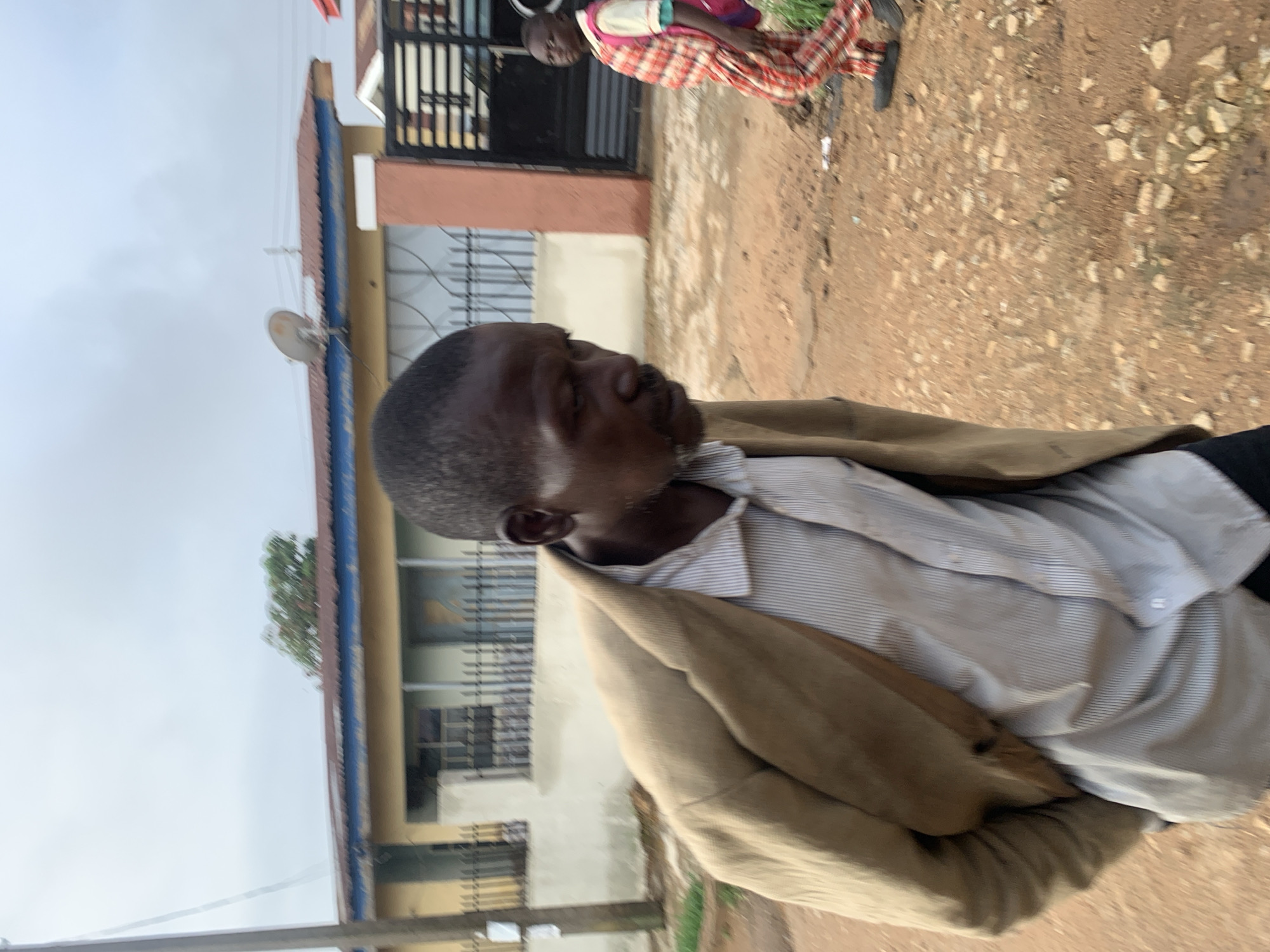 This image depicts a street scene in what appears to be Jos, Nigeria, captured on an overcast day. The primary subject is a dark-skinned man positioned prominently in the foreground, seen from the chest up. He has short dark hair and a beard or stubble, and his gaze is directed slightly downwards and to his left. He is wearing a light-colored, vertically striped button-up shirt beneath a tan or light brown jacket.
Behind the man, the ground transitions from a paved-like surface to an unpaved area strewn with dirt and gravel. In the mid-ground, further back and to the right, another individual, possibly a child or smaller adult, is visible. This person is dressed in a red and white plaid or checkered garment and is standing near a building.
The background features various buildings with different architectural styles. One structure displays reddish-brown walls and has a black metal gate or fence in front. Another building segment shows a distinct blue trim and has a small white satellite dish or antenna mounted on a pole. The sky above is cloudy and uniformly grey, suggesting an overcast daytime. The scene appears to be a candid snapshot of daily life in an urban or semi-urban setting, with no specific event unfolding. There is no visible text in the image.
