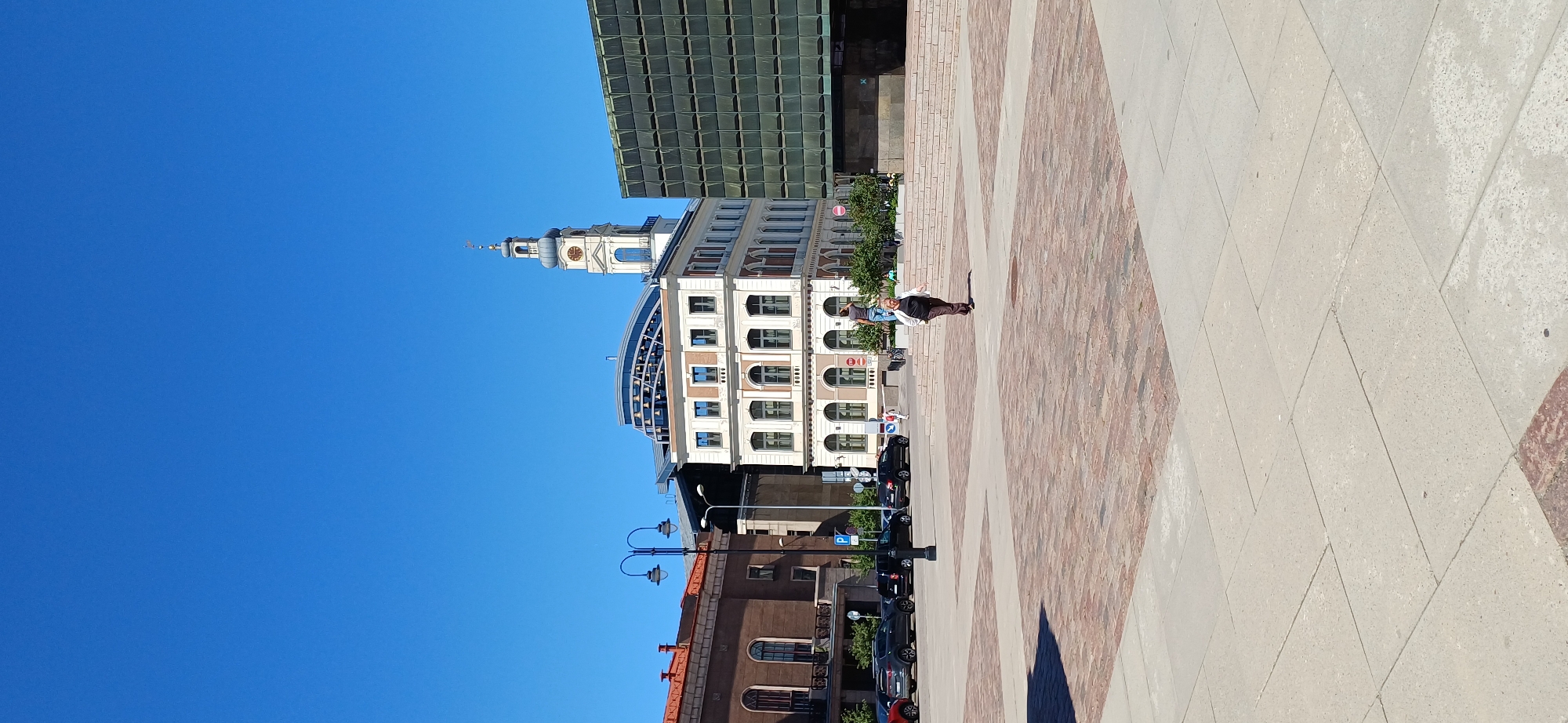 In a city square, under a clear blue sky, a lone person walks across a paved area. The square is bordered by buildings, one a classical building with arched windows and a clock tower, another a modern building with a glass facade. A few cars are parked along the street in the background, and a pair of ornate street lamps stands near a building. The ground is paved with light-colored rectangular stones, with sections of red brick paving. The person walking is a woman with dark hair, wearing a dark jacket over a white top and dark pants. She appears to be walking towards the viewer, with her head tilted down slightly.