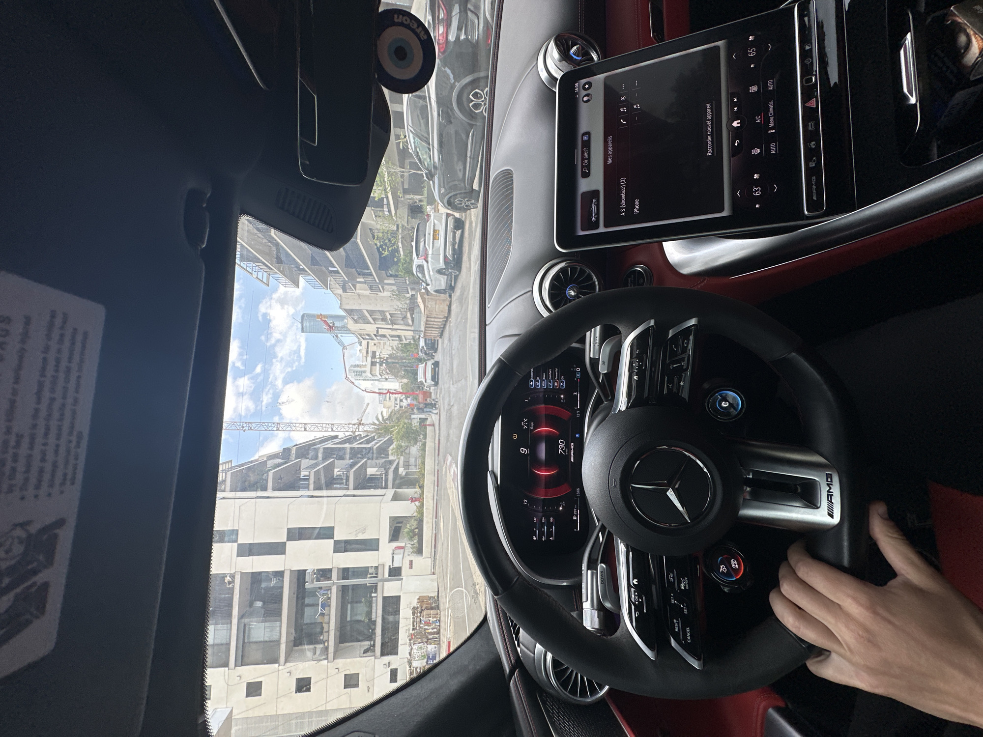 The image shows the interior of a Mercedes-Benz AMG, likely a 2023 SL-Class, with the dashboard, steering wheel, and digital instrument cluster in focus. The steering wheel is black with silver accents and an AMG logo. The instrument cluster displays information like the current speed (9 km/h) and RPM (730). A hand is visible on the right side of the steering wheel.
The central infotainment screen is displaying "Mes appareils" and lists an iPhone as a connected device. Climate control settings are visible below the screen, with temperatures of 63 and 65 degrees displayed.
Through the windshield, a cityscape is visible. Buildings under construction are evident, as well as multiple cars parked along the street. Based on architectural styles and ongoing construction, the location is Tel Aviv, Israel.