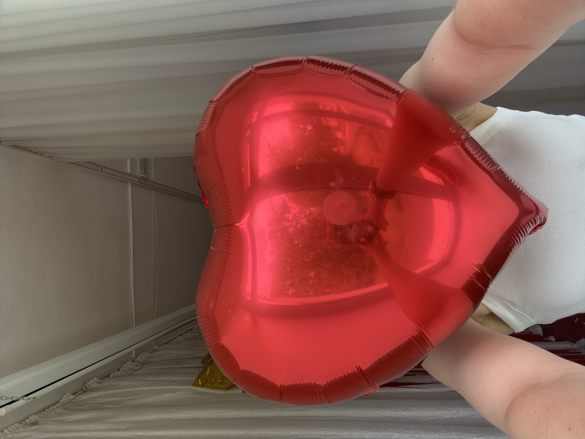 A person holds a red, heart-shaped balloon indoors. The location is not identifiable as Chaikovsky, Russia, or any other specific location based on the visual information provided. The person wears a white shirt and is seen holding the balloon in their arm. The background appears to be a room with neutral-colored walls and a ceiling with evenly spaced panels. There's a window with white blinds.