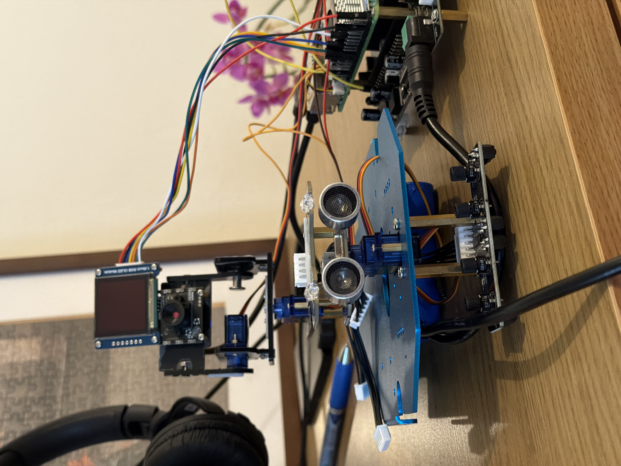 The image depicts an electronic assembly, seemingly a prototype robot or automated system, situated on a wooden surface. The assembly includes components such as a small camera module with an accompanying display screen, ultrasonic sensors, circuit boards stacked vertically, and various connecting wires and cables. The components are mounted on a blue chassis. The background includes a glimpse of a framed picture and a headphone. The location is unknown and there is no indication of this picture being located in Givatayim, Israel.