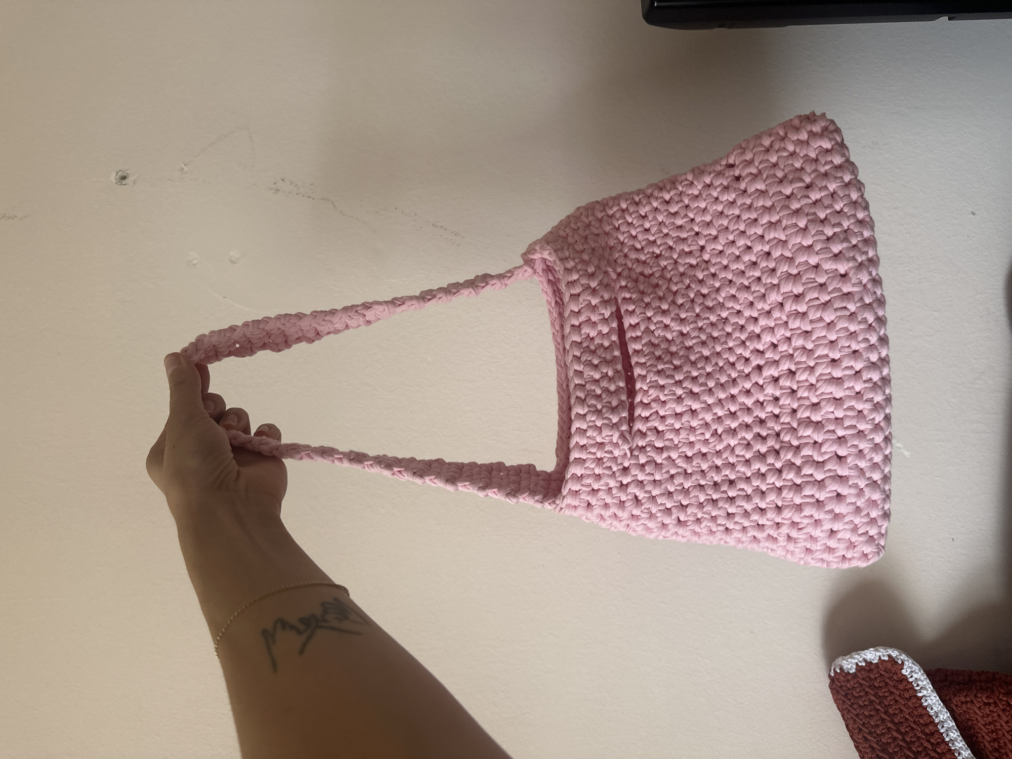 A person's left forearm, adorned with a tattoo and a delicate bracelet, is visible, holding a pink crocheted shoulder bag. The bag, with its braided strap and textured body, hangs against a pale pink wall that has visible imperfections and a small dark mark near the top right. To the left and partially out of frame, a portion of a brown crocheted item with a white trim is also visible. The scene appears to be indoors, possibly a room in Gyumri, Armenia, given the context.