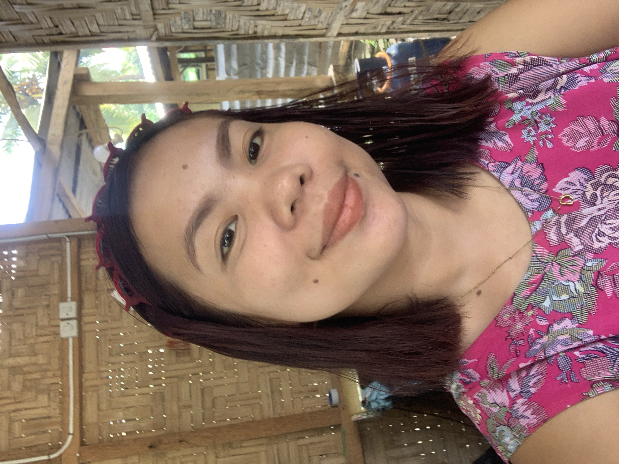 A young woman with dark hair, wearing a pink floral dress and a red headband, smiles at the camera in an outdoor setting with a bamboo wall and roof visible in the background. The location is identified as Kiblawan, Philippines.