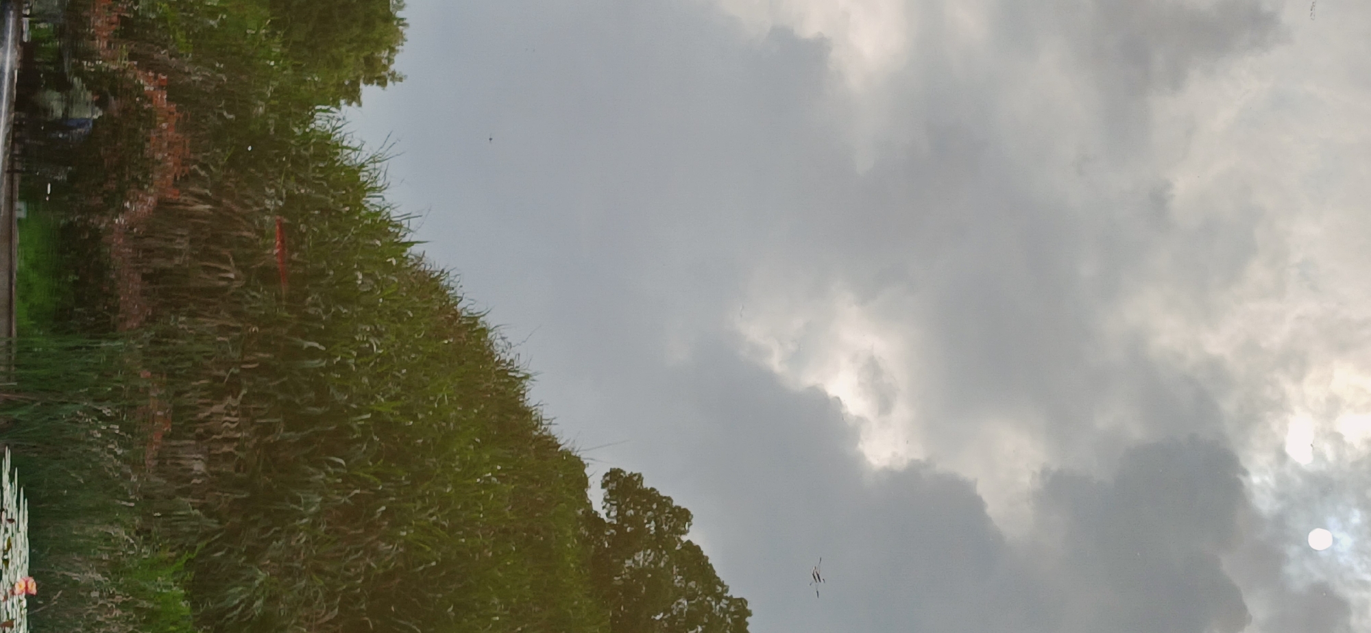 The image shows a reflection of a cloudy sky and green vegetation, likely trees and reeds, in a body of water. The sun is visible through the clouds. A red flag is also visible. The reflection also shows what appears to be part of a structure, possibly a building. The location is specified as Chongqing High-tech Zone, China, but no specific details corroborate this.