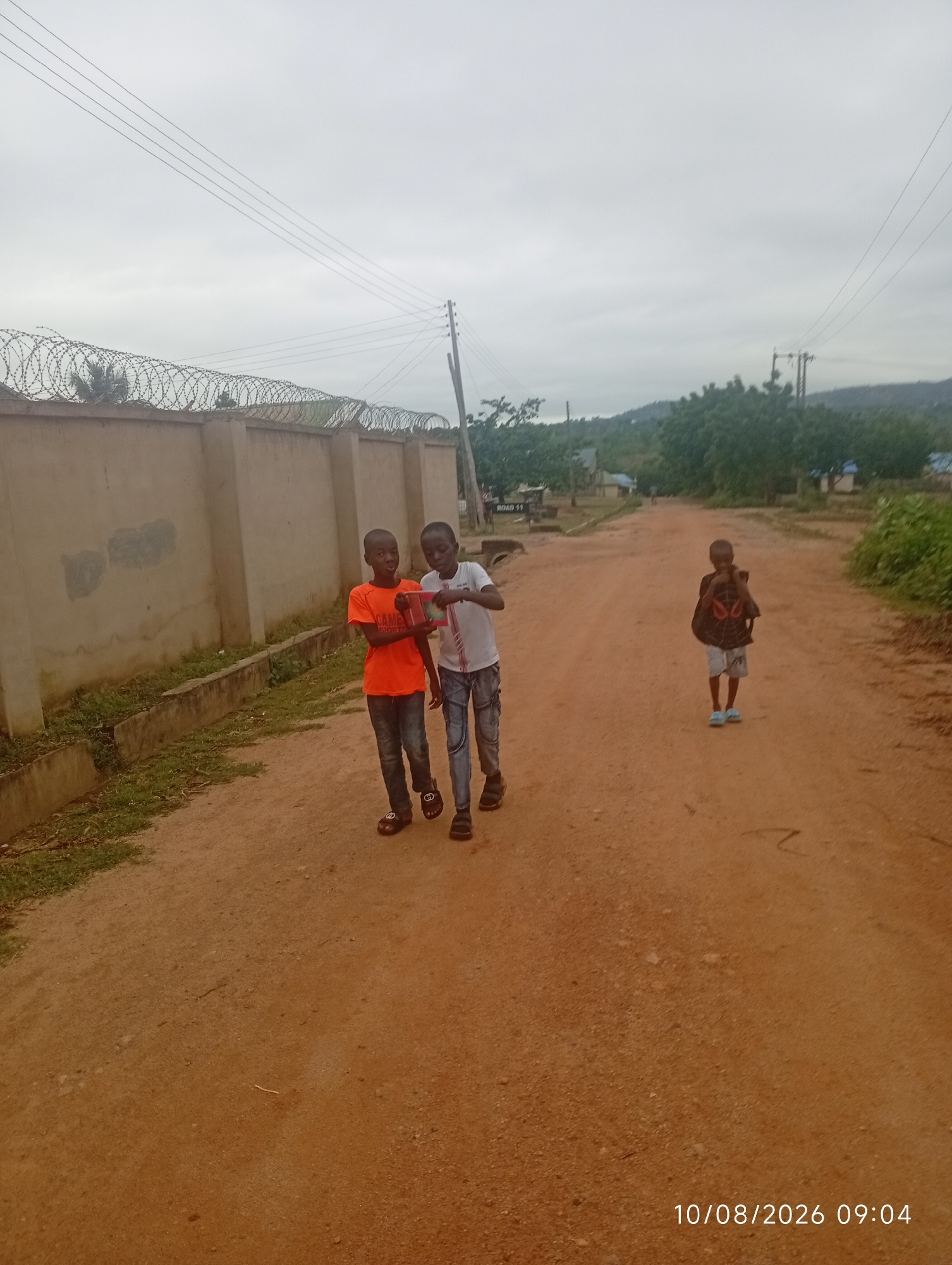 Three young boys walk on a dirt road in an outdoor, rural setting. The boy on the left wears an orange t-shirt and blue jeans, while the boy in the middle wears a white t-shirt and blue jeans. The boy on the right wears a black shirt with a red spider-like emblem and light-colored shorts. A beige wall topped with barbed wire runs along the left side of the road. Trees and hills are visible in the background. The sky is overcast. A timestamp in the bottom right corner reads "10/08/2026 09:04".