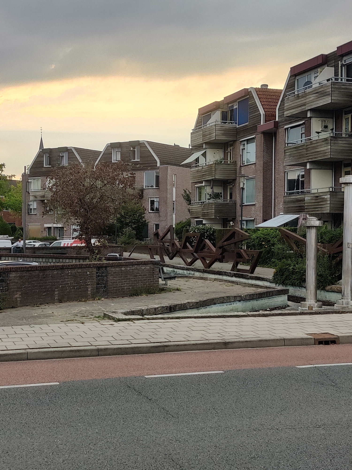 The image shows an overcast sky over a row of multi-story apartment buildings in Winterswijk, Netherlands. In the foreground, a street with a red-colored lane is visible, separated by a curb from a paved sidewalk. Behind the sidewalk, there is a low brick wall surrounding a small courtyard or garden area. Within this area, a modern abstract sculpture made of dark metal is prominently featured. Adjacent to the sculpture are lush green bushes and two ornate cylindrical pillars. Beyond the courtyard, cars are parked in front of the apartment buildings, some of which have balconies with wooden cladding. A bare tree with reddish-brown leaves stands in the middle ground, to the left of the sculpture. The buildings appear to be residential, with multiple windows and balconies. The overall scene suggests an urban environment with a mix of residential architecture and public art.