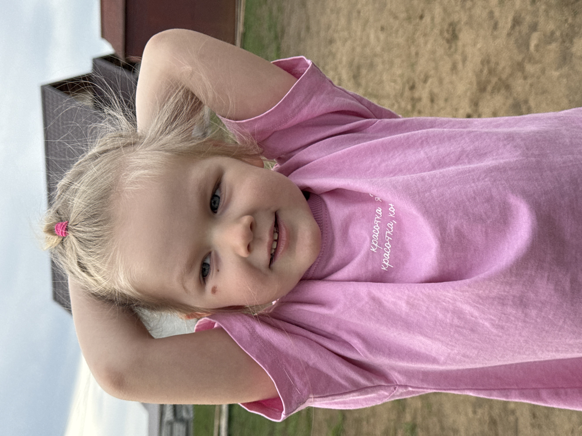 A young, fair-skinned child with blonde hair is the main subject of the image. The child is wearing a pink T-shirt with white text. The child is lying on their back with their arms raised, hands behind their head, and is looking directly at the camera with a slight smile.
The background shows a glimpse of the outdoor environment, including a wooden building or structure, suggesting a rural setting. The ground is covered in grass or dirt.
The location is ферма, Russia.