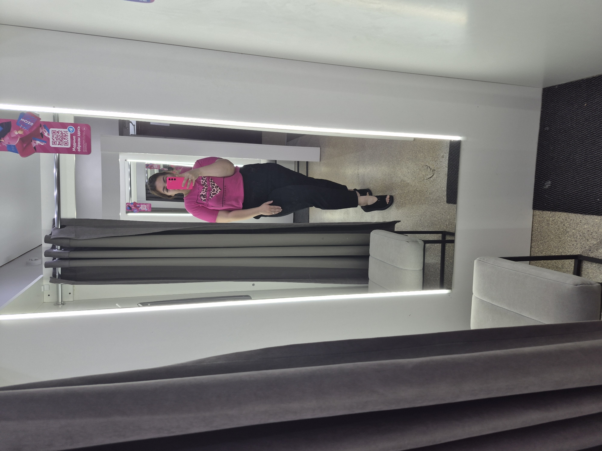 A woman is taking a mirror selfie in a fitting room. She is wearing a pink t-shirt with a leopard print heart and black pants. She is standing in front of a large mirror, with a gray curtain to her left and a white wall with a light strip to her right. There is a gray bench with a black metal frame in the foreground. The floor is carpeted. A QR code poster is visible on the wall in the mirror's reflection. The image appears to have been taken in a retail store, possibly in Chaikovsky, Russia, given the Cyrillic text on the poster.