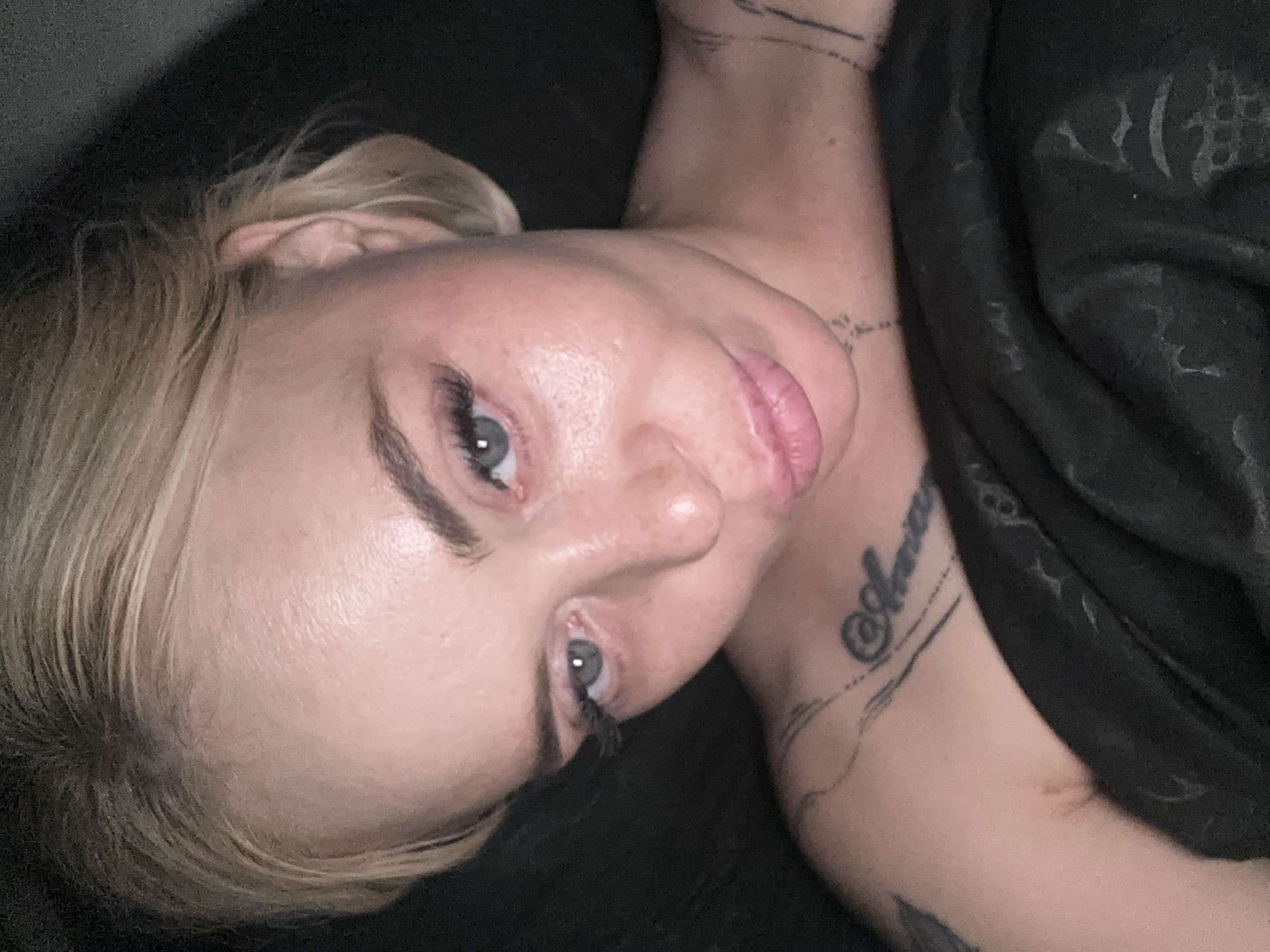 A close-up, high-angle, indoor shot features a young, blonde-haired woman with fair skin and light blue eyes. Her hair is pulled back loosely, revealing her forehead, ears, and the side of her face. Her eyes are looking slightly to the left of the camera, and she has on false eyelashes and light makeup. Her lips are slightly parted and appear to be a light pink color. Tattoos are visible on her upper left shoulder and across her collarbone. She is covered from the chest down with a dark, textured fabric, possibly a blanket or clothing. The background is dark and out of focus. The lighting is dim, casting shadows on her face and hair.