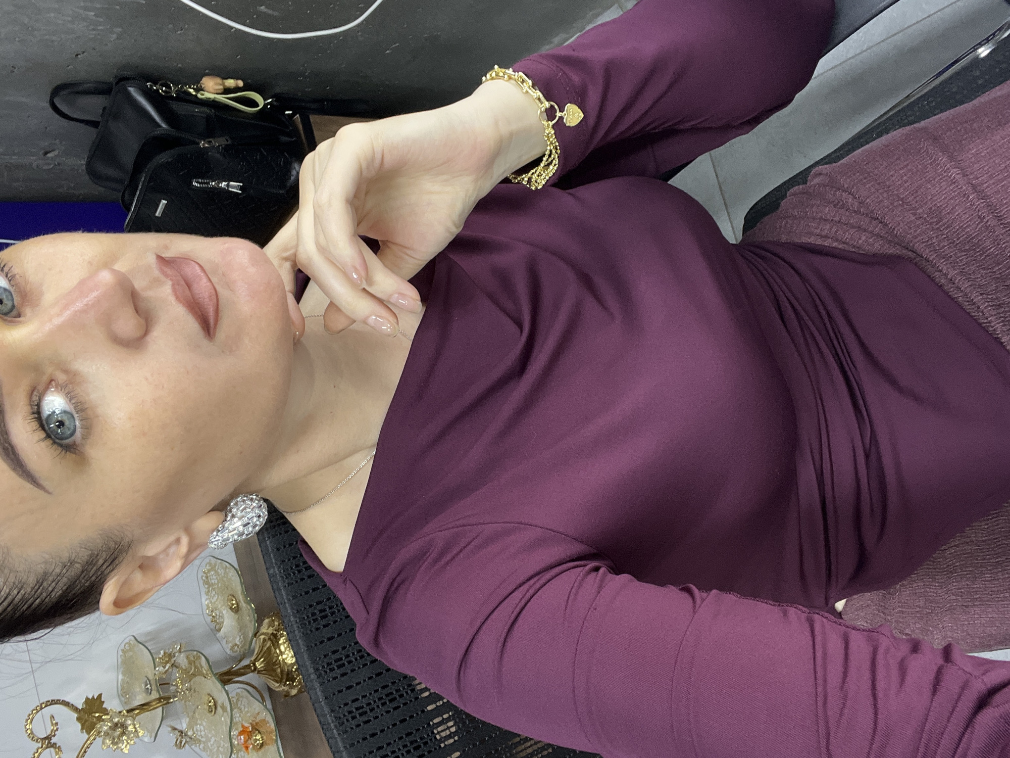 In the city of Shahrixon, Uzbekistan, a medium shot captures a woman seated in a black chair, her body angled towards the camera. She wears a deep plum-colored long-sleeved top with a draped neckline and a textured plum-colored skirt. Her left hand rests on her chin, showcasing a gold bracelet with a heart-shaped charm and neatly manicured nails. She sports large, diamond-encrusted earrings and a delicate silver necklace. Her makeup includes defined eyebrows, subtle eyeshadow, and a nude lipstick. The background reveals a gold ornate multi-tiered display stand with decorative plates on a wooden surface, a black leather backpack with a lime green keychain and a small teddy bear charm, and a grey wall.