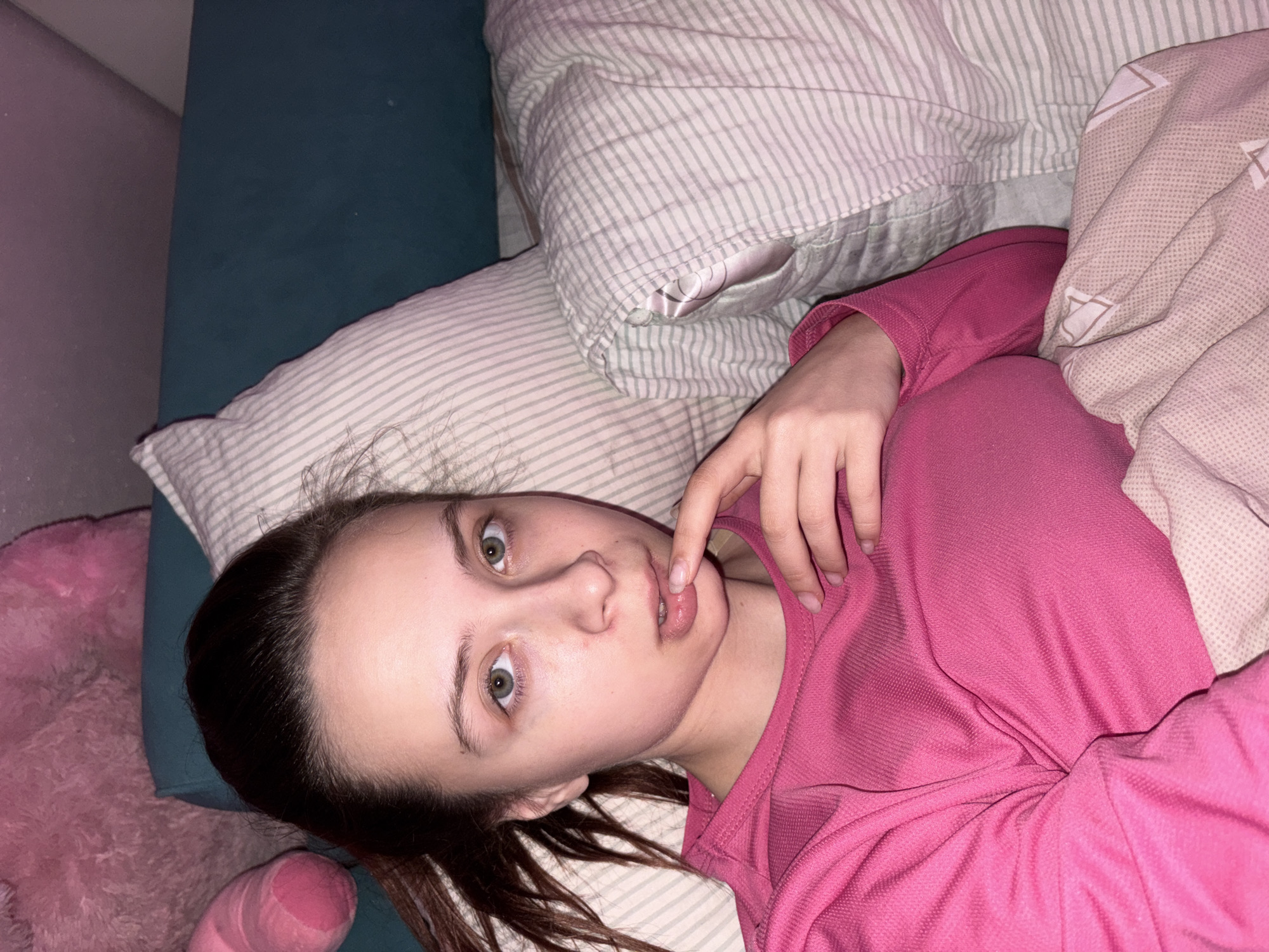 A young female individual with fair skin and light green eyes is lying in a bed. She is wearing a pink long-sleeved shirt. Her dark hair is loose around her head and partially under her. Her left hand is raised, with a finger touching her lower lip. The bed has white and grey striped pillows and a tan and white patterned blanket. In the background to the left, part of a pink stuffed animal and a dark teal headboard are visible. The location is likely an indoor residential space. No definitive evidence can be derived to determine if the image was taken in the city of Прикамский, Russia.