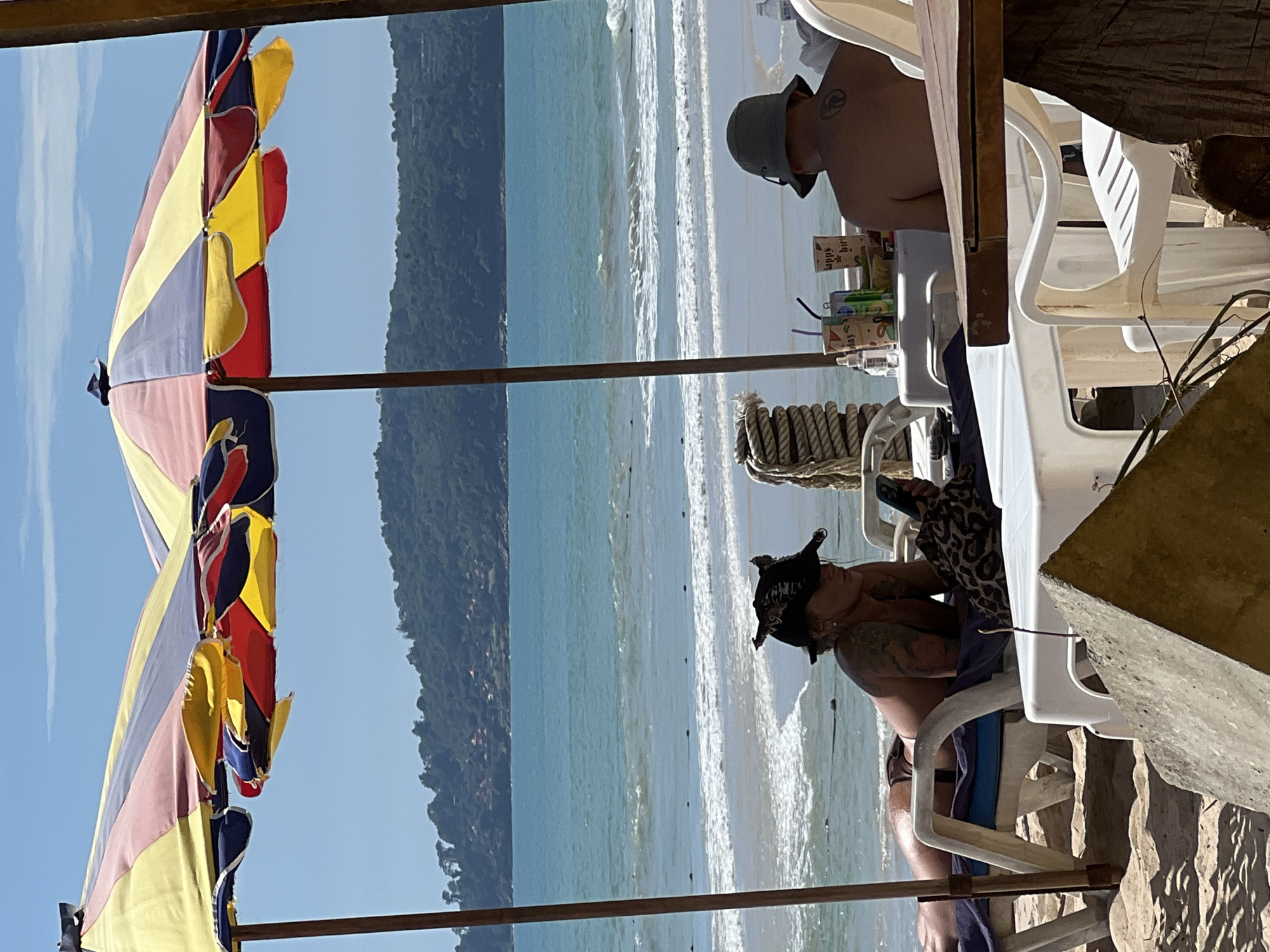 The image captures a scene at a beach in Unknown, Unknown. Two people are visible under large, colorful beach umbrellas. On the left, a woman with tattoos is lying on a lounge chair, wearing a black hat and holding a cell phone. She is facing towards the ocean. On the right, a person wearing a green hat is seated, facing away from the camera, with a tattoo on their back. In front of the seated person, there is a white table with cups and drinks. The ocean with gentle waves is visible in the background, with a tree-covered hill in the distance under a clear blue sky. The foreground shows sandy ground and a portion of a wooden structure.