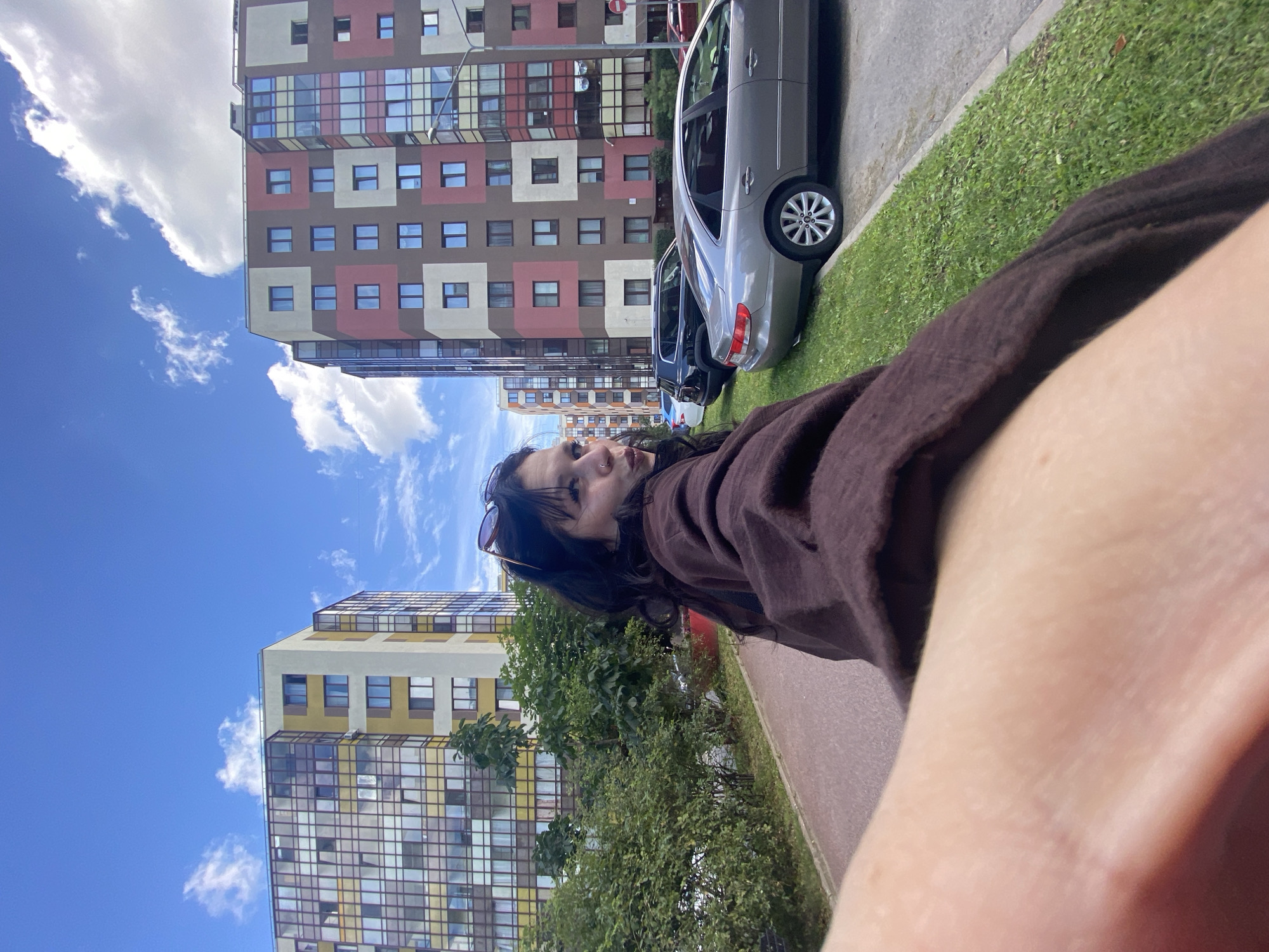 An outdoor, medium-distance, eye-level, slightly low-angle shot captures a young woman with dark hair and sunglasses on her head, looking over her shoulder at the camera while holding the camera with her right arm. She is wearing a loose, dark brown hooded sweatshirt. The woman is standing on a red paved walkway, facing a row of parked cars and a cluster of tall, modern apartment buildings in Kudrovo, Russia. The buildings are colorful, with alternating panels of yellow, white, brown, and pink. A strip of green grass with a curb separates the walkway from the street where several cars are parked, including a silver sedan and a black SUV. The sky is blue with scattered white clouds.