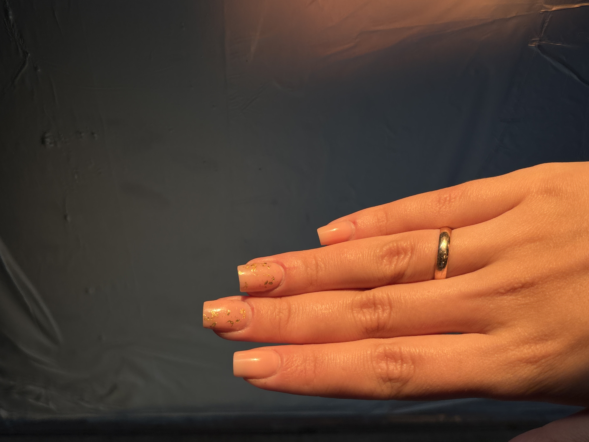 A close-up, eye-level shot shows a left hand with manicured nails against a dark, textured background. The background appears to be a draped material with wrinkles and subtle highlights from lighting, suggesting an indoor setting. The hand is illuminated by warm, directional light, casting shadows and highlighting skin texture. The fingernails are painted a pale peach color, with the middle and ring fingers adorned with gold foil accents. A thin, gold band is visible on the ring finger. The image composition focuses on the detail of the hand and its adornments, with the background serving as a neutral, textural element. The location in the city of Inskoy, Russia is not determinable from the visual information present in the image.