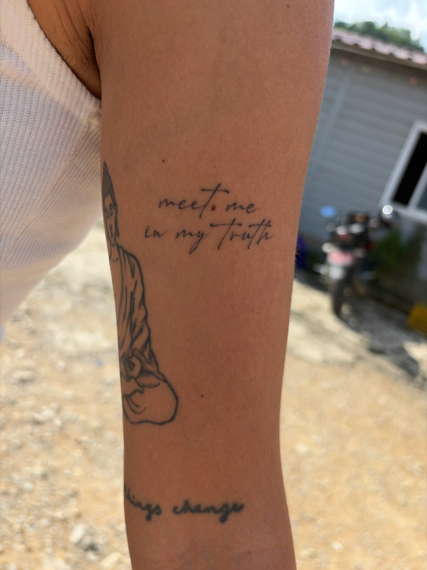 The image shows a close-up of a person's forearm with a tattoo of a seated figure, possibly a Buddha, and text in cursive script. The text reads, "meet me in my truth" and below that, partially visible, "things change". The skin is tanned. The background is blurred but suggests an outdoor environment with a gravelly or sandy ground. In the far background, a building and a motorcycle are faintly visible, which could indicate a location in or near Vyas (Damauli), Nepal, a region known for its scenic beauty and connection to Buddhist culture. The lighting suggests daylight.