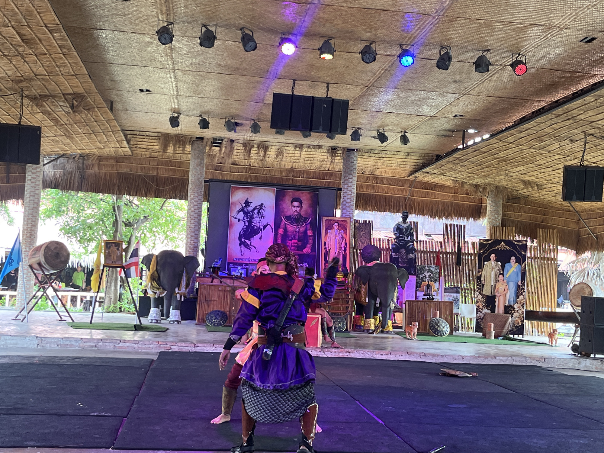 In Ayothaya Town Municipality, Thailand, an outdoor stage performance is underway beneath a thatched roof structure. Two performers, dressed in traditional vibrant purple and yellow attire with swords, engage in a scene of swordplay on a black mat. Behind them, a large screen displays images of regal figures. To the left, a large drum on a wooden stand and an elephant adorned with gold fabric stand as part of the performance's backdrop. Further to the right, another elephant and a display of portraits of Thai royalty, including what appears to be King Bhumibol Adulyadej and Queen Sirikit, are visible. The environment is set up for a cultural event, with decorative elements and lighting enhancing the stage.