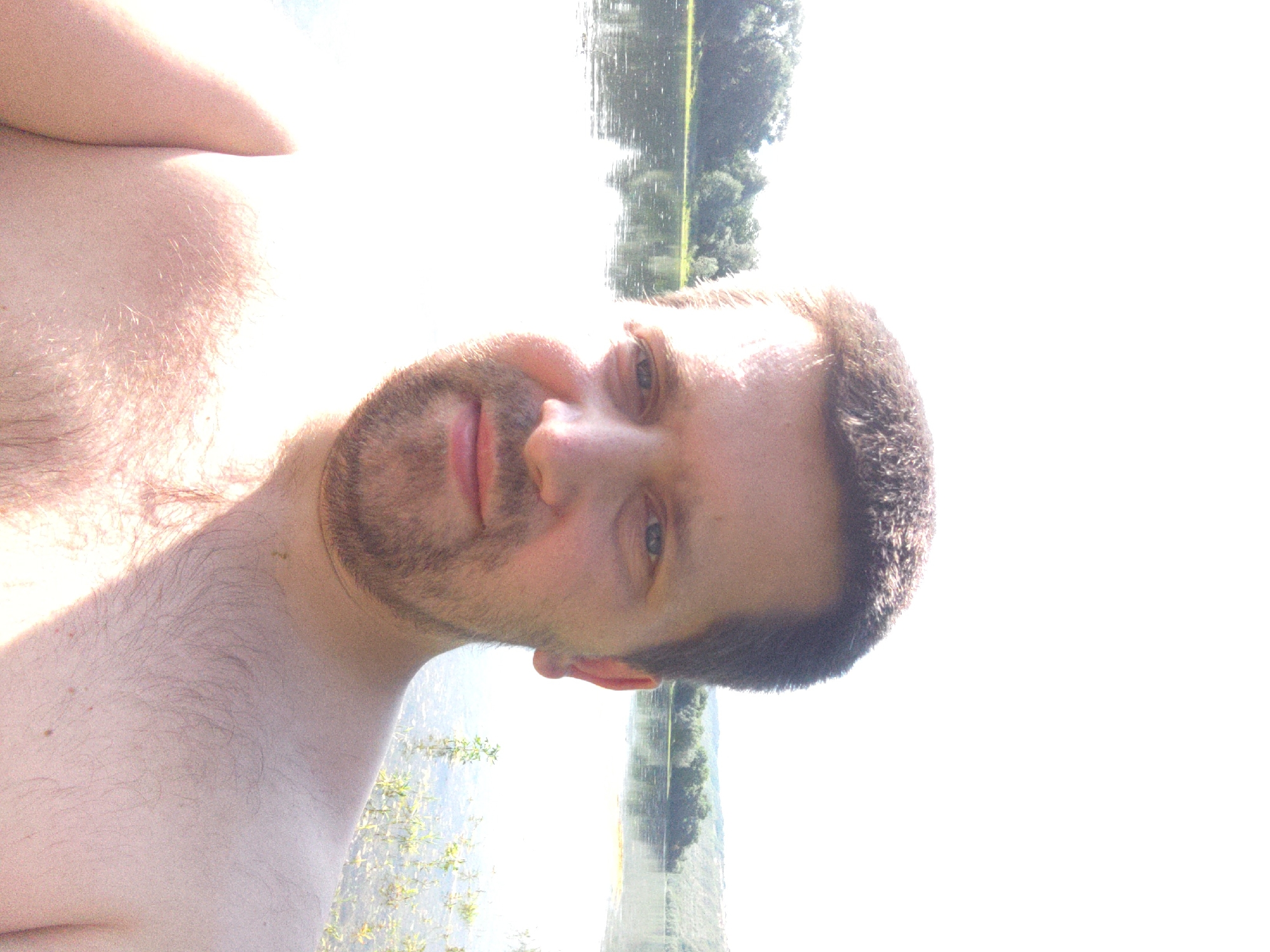A man is taking a selfie while in the water, possibly swimming or wading. The image is taken from a low angle, with the man's chest, shoulder, and part of his face visible. He has fair skin, light brown hair, and a beard. His eyes are open and looking towards the camera. The background shows a body of water with reflections of trees and the sky, suggesting a sunny day. The man's expression appears neutral or relaxed. The scene is outdoors, with vegetation visible along the water's edge. The photograph appears to be taken during daylight hours, likely in the late morning or afternoon due to the brightness of the sky and the shadows cast. The provided location context of Zalishchyky, Ukraine, suggests a rural or natural setting. There is no visible text in the image.