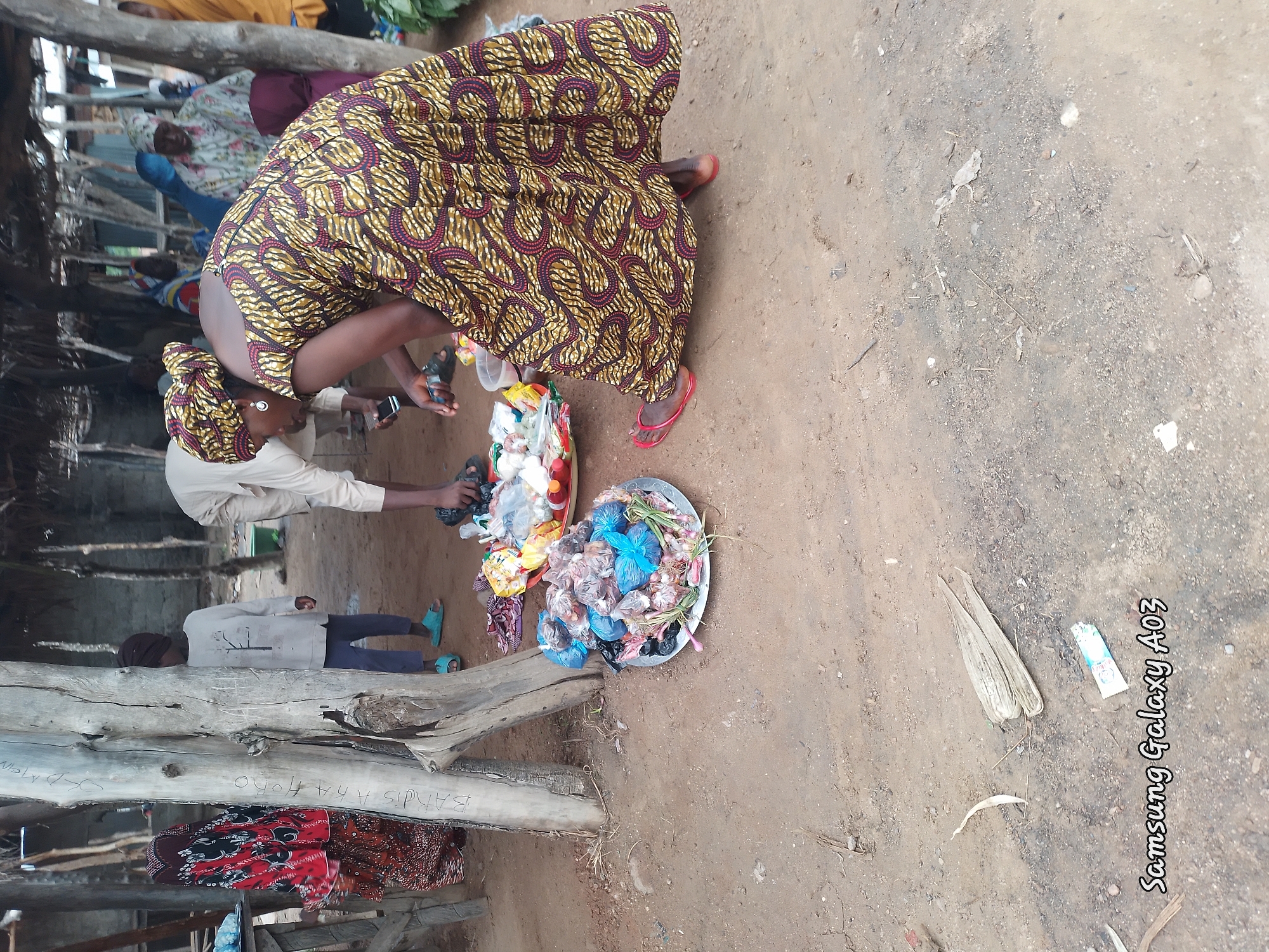 In Doka-Tofa, Nigeria, this outdoor scene captures a vibrant market setting. A woman, adorned in a striking yellow and maroon patterned dress and headwrap, is engaged in commerce. She is bent over, interacting with goods displayed on a metal platter, which appears to contain a variety of items, possibly food or spices, wrapped in plastic bags. Another individual, wearing a light-colored garment and head covering, stands behind her, also appearing to be involved in the transaction or browsing.
In the background, several other individuals are visible under a thatched roof structure, suggesting a marketplace with multiple vendors and shoppers. A child in a gray sweater and blue pants is also present, standing near a wooden post. The ground is covered in dry earth, typical of a rural or semi-urban environment. Two metal platters are on the ground, one containing an assortment of items in plastic bags, and the other holding what seems to be vegetables or herbs. The overall atmosphere is that of an active local market.