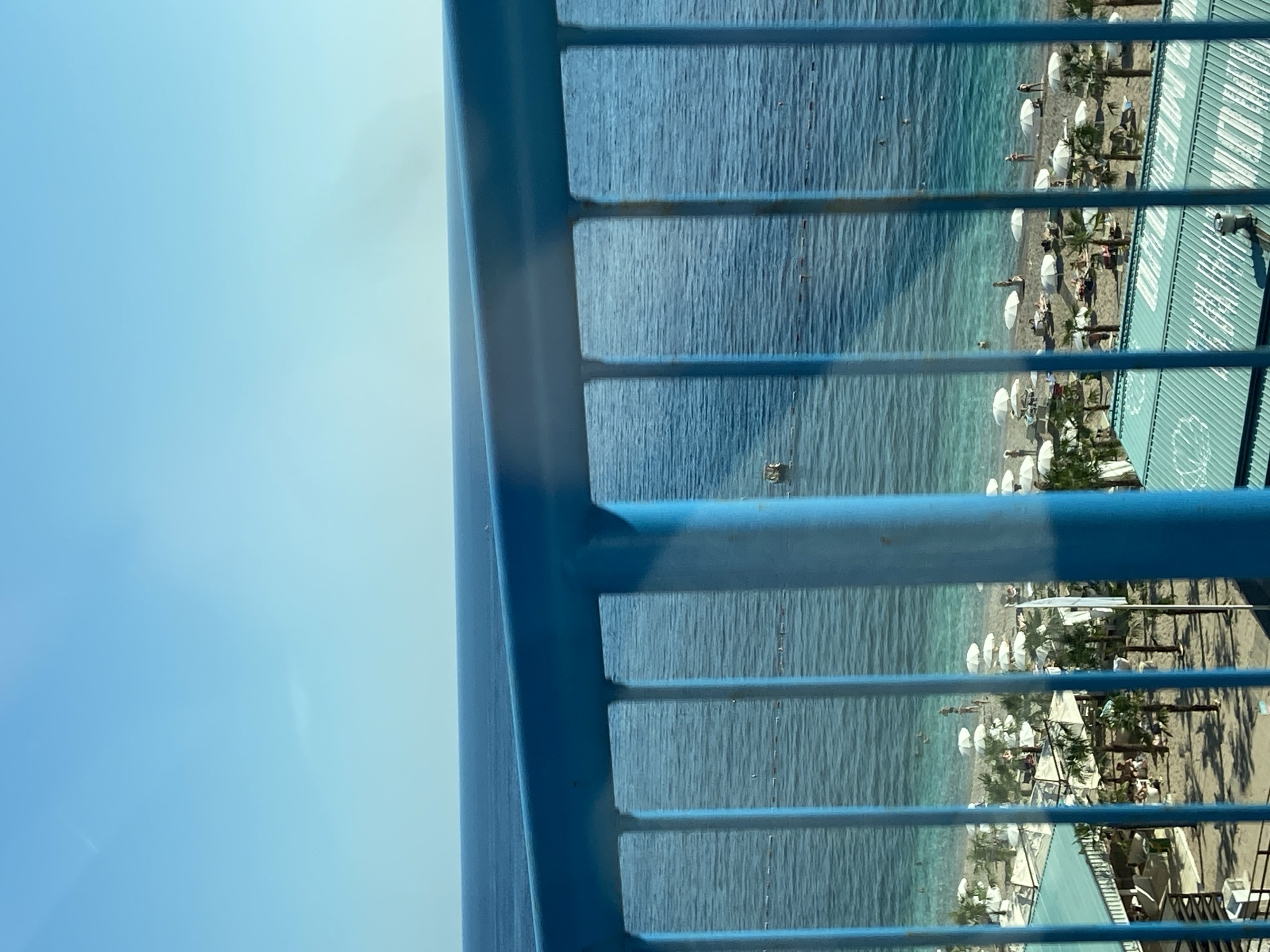 The image shows a view of a beach in Rafailovići, Montenegro, seen through a blue metal railing. The sea is visible, with people swimming. The beach is lined with white umbrellas and palm trees. Buildings are on the edge of the beach. The image is oriented sideways.