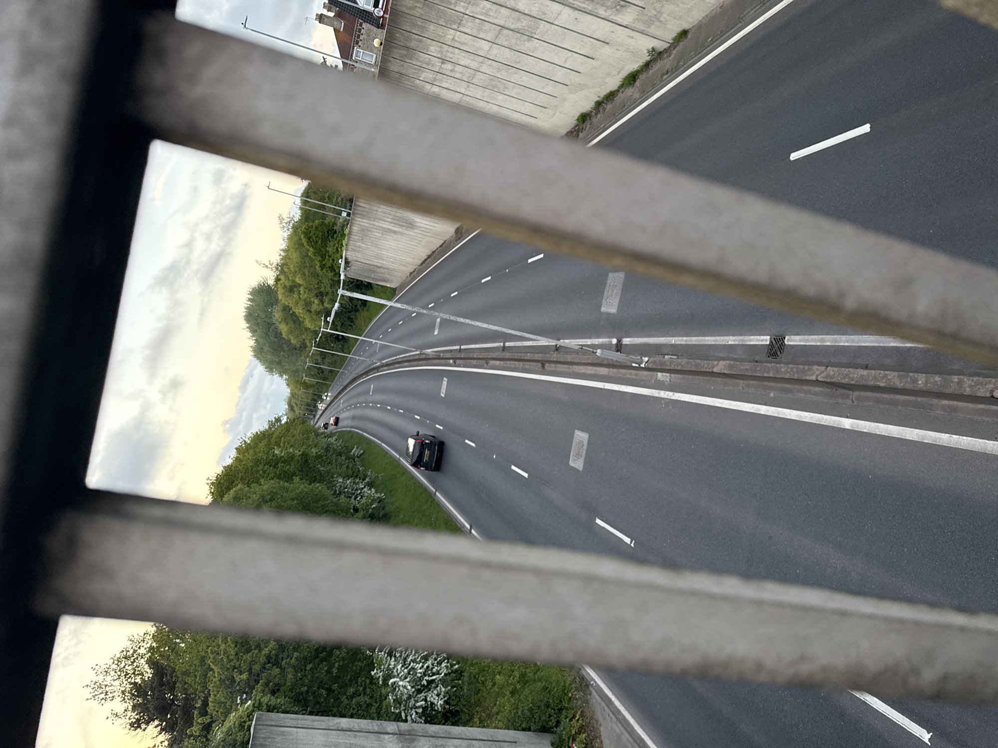 The image shows a view of a highway through a metal barrier. The highway has multiple lanes and a concrete barrier on one side. A few cars are visible on the highway. Trees and vegetation are on both sides of the highway. In the background, buildings are visible. The highway appears to be curving. The image was taken from Great Yarmouth, United Kingdom.