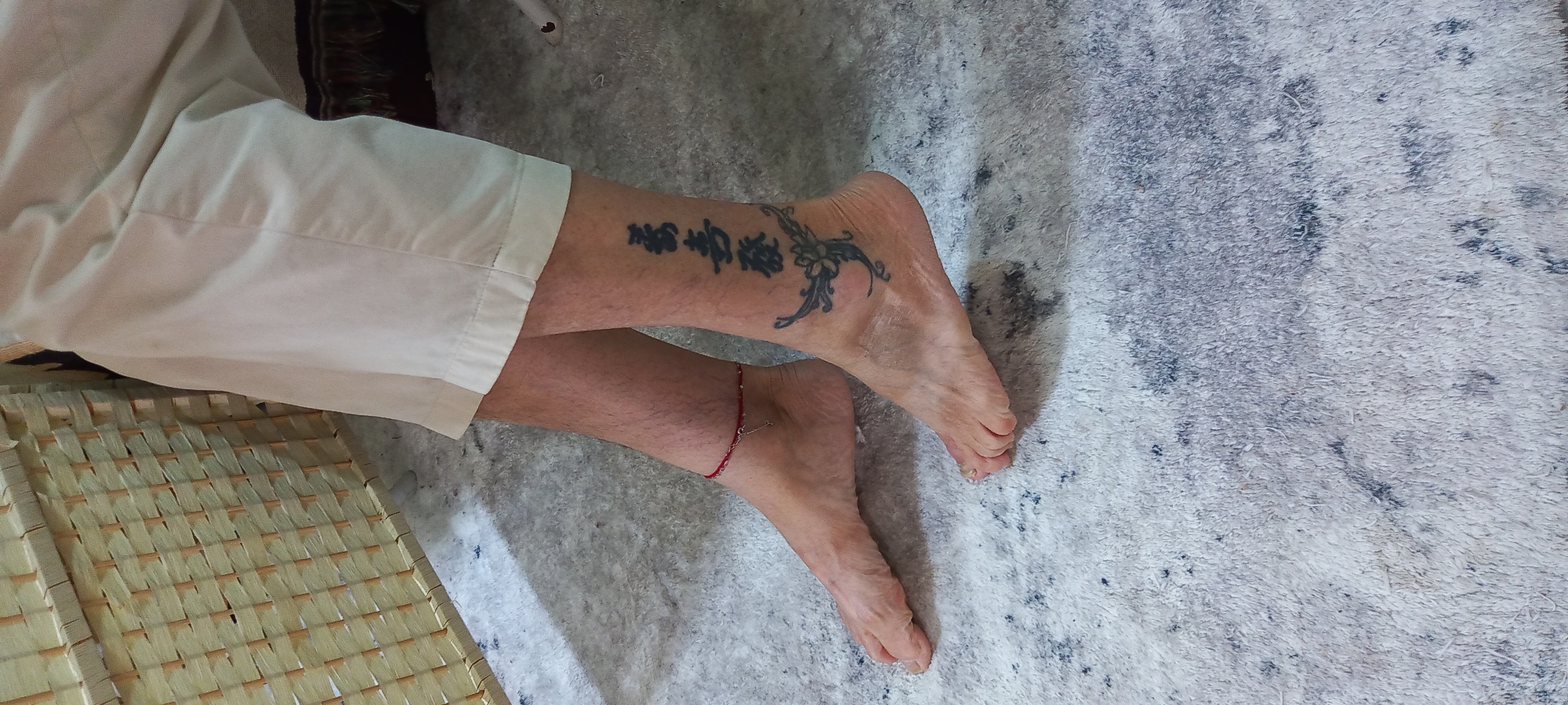 This image, taken in Baan Chaloklum, Thailand, captures a close-up view of a person's lower legs and bare feet. The individual is wearing light-colored, possibly off-white, casual pants, with the fabric visible above the ankles. A portion of a light-colored, woven mat or furniture piece with a crisscross pattern is seen on the bottom left.
The person's right leg displays two prominent tattoos. On the upper calf, there are two distinct East Asian characters, which translate to "喜樂" (xǐlè), meaning "joy" or "happiness." Adjacent to these characters, extending towards the ankle, is a black, ornamental tattoo featuring a stylized floral or vine design with leaves. A thin red string anklet, adorned with a small metallic charm, is visible on the left ankle.
The legs and feet rest on a soft, light-colored carpet or rug. The carpet features an irregular, mottled pattern in shades of white, grey, and black, suggesting a textured, possibly shag-pile surface. The overall lighting indicates an indoor setting during the daytime. The scene portrays a casual and relaxed atmosphere, with the person seemingly at rest.