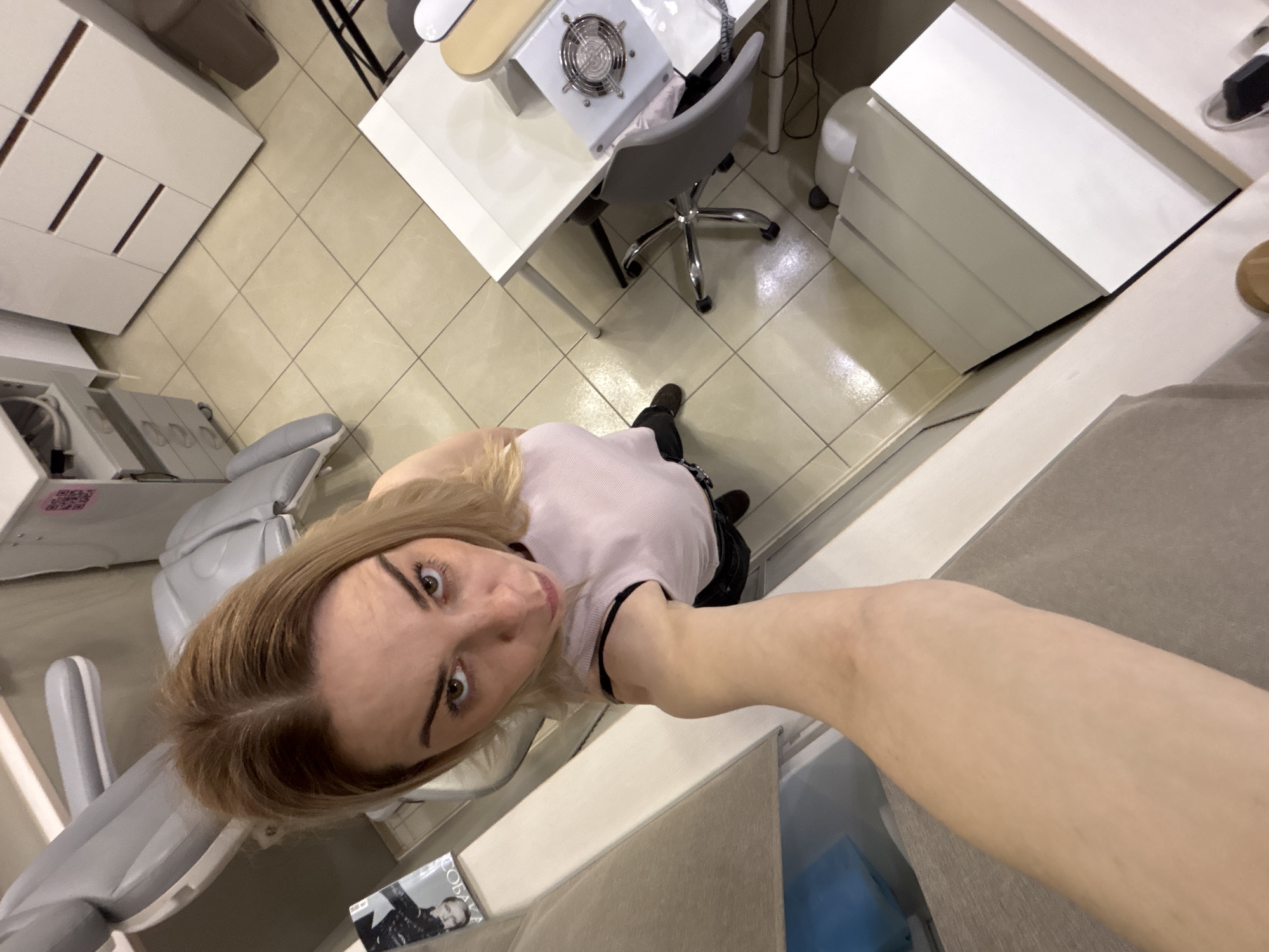 A high-angle, point-of-view shot shows a woman with blonde hair and a light pink shirt taking a selfie. She is indoors, possibly in a beauty salon or clinic. The environment includes a light-grey treatment chair, a white table with a manicure dust collector on it, a white storage cabinet, and tiled flooring. A magazine is visible on the chair. The location is potentially in Vsevolozhsk, Russia.