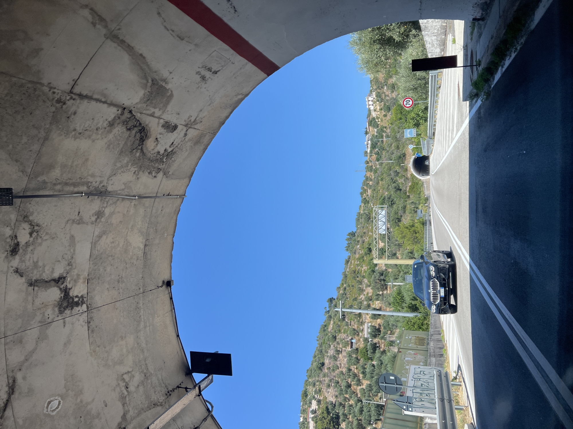 This media file shows a view from inside a tunnel, looking out towards a bright blue sky and a sunlit hillside. The tunnel opening is on the left, revealing a rough, textured concrete ceiling with some staining and a red stripe at the top. A dark rectangular object is mounted on the tunnel wall.
The scene outside the tunnel is a road with a black car driving on it. The road is bordered by a metal guardrail and white lane markings. The hillside is covered in lush green vegetation, including olive trees, and dotted with buildings. A speed limit sign showing "70" is visible on the right side of the road. Further down the road, another tunnel entrance is visible. The overall impression is a sunny day in a scenic, possibly coastal, Italian location like Mattinata, Italy, given the context. The time of day appears to be daytime, and the weather is clear and sunny.