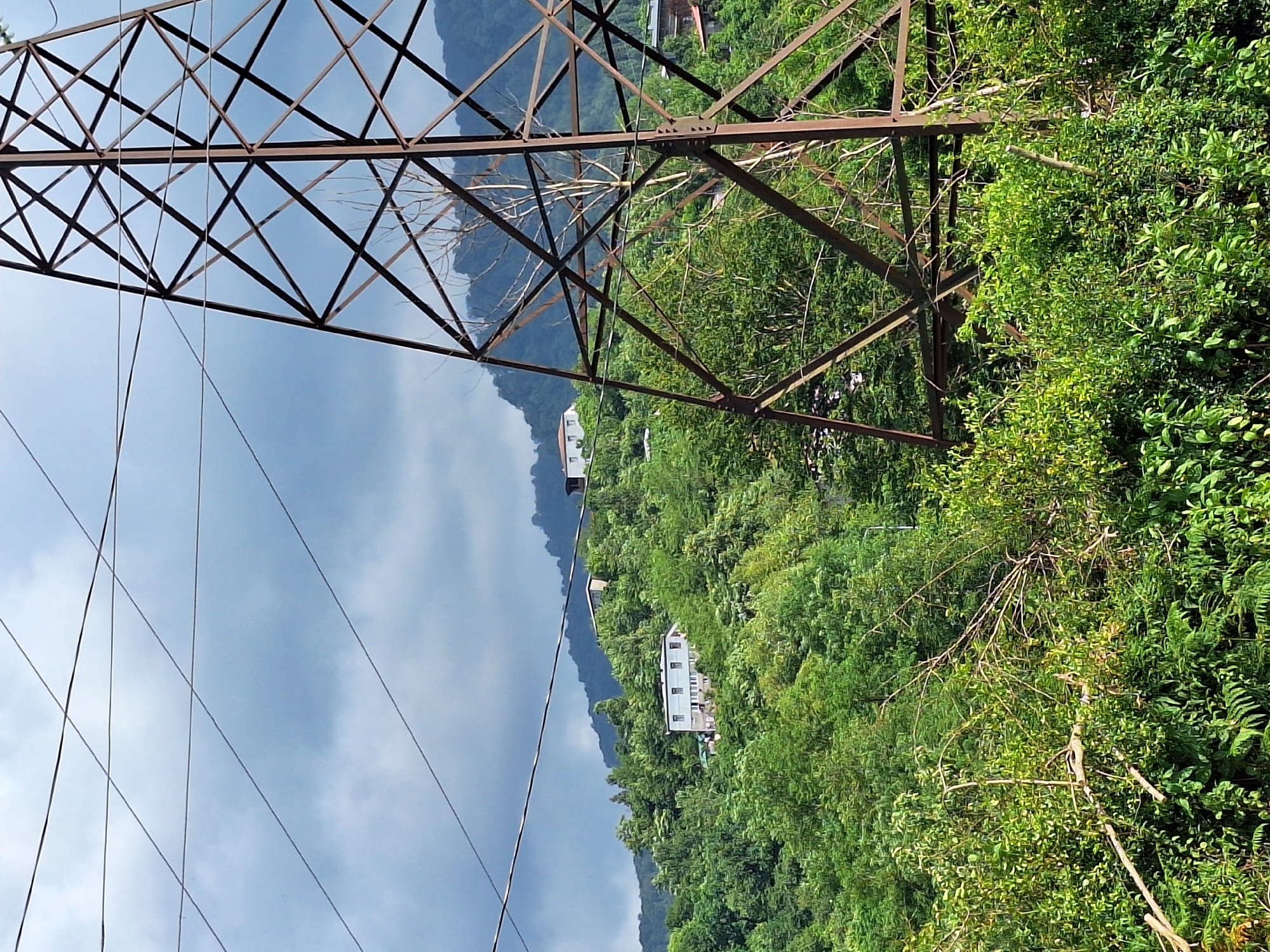 A wide-angle, slightly low-angle shot captures a mountainous landscape near Kobuleti, Georgia. Dominating the foreground is a large, rusty metal electricity pylon, its lattice structure visible against a cloudy, grey sky. Several high-voltage power lines traverse the frame, creating a crisscrossing pattern against the sky. Beyond the pylon, a lush green hillside is dotted with several residential buildings, varying in size and design. The vegetation is dense, with a mix of trees and smaller plants covering the slopes. The sky is overcast with dark, heavy clouds, suggesting impending rain or a generally gloomy day. The overall impression is one of a rural or semi-rural area with essential infrastructure, likely serving a residential community on the mountainside.