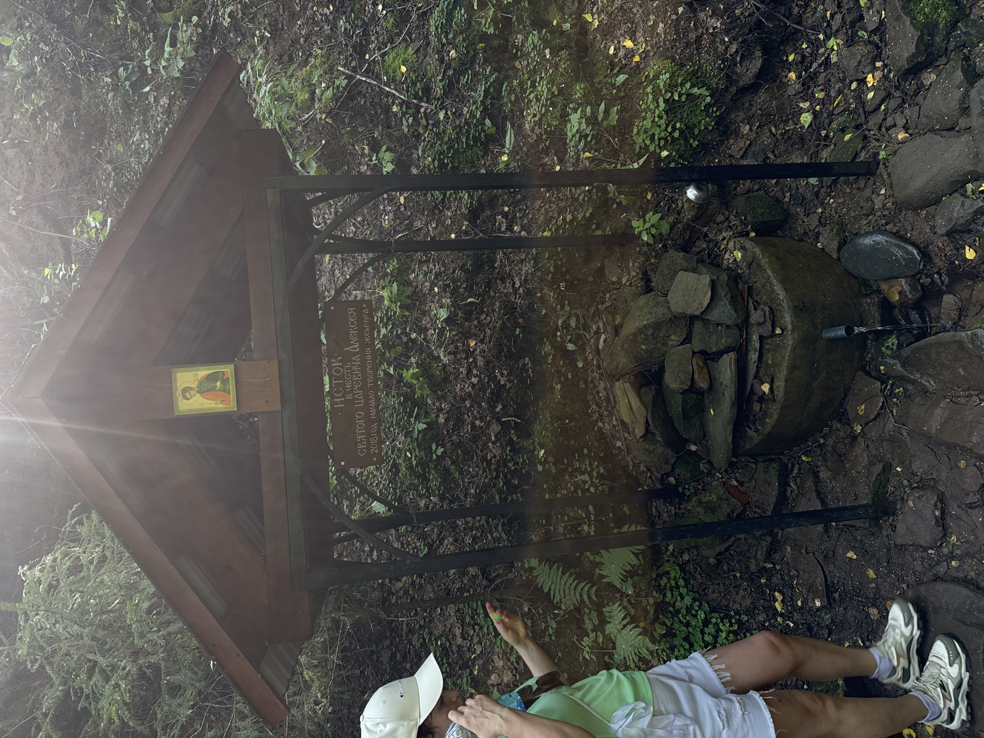 An outdoor scene depicts a wooden structure with a sloped roof, housing an icon of a religious figure. Below this structure is a stone basin with rocks piled on top, and a spigot from which water is flowing. A person wearing a white cap, green t-shirt, and white shorts stands to the left, holding a clear bottle and a small green object. They are wearing white and gray sneakers. The environment appears to be a forest with trees and dense foliage. The sign on the structure indicates that this is a spring (источник) in honor of Saint Tsarevich Alexei. The inscription also states that the creation (начало творения) began in 2018. This location is identified as a spring in Izhevsk, Russia.