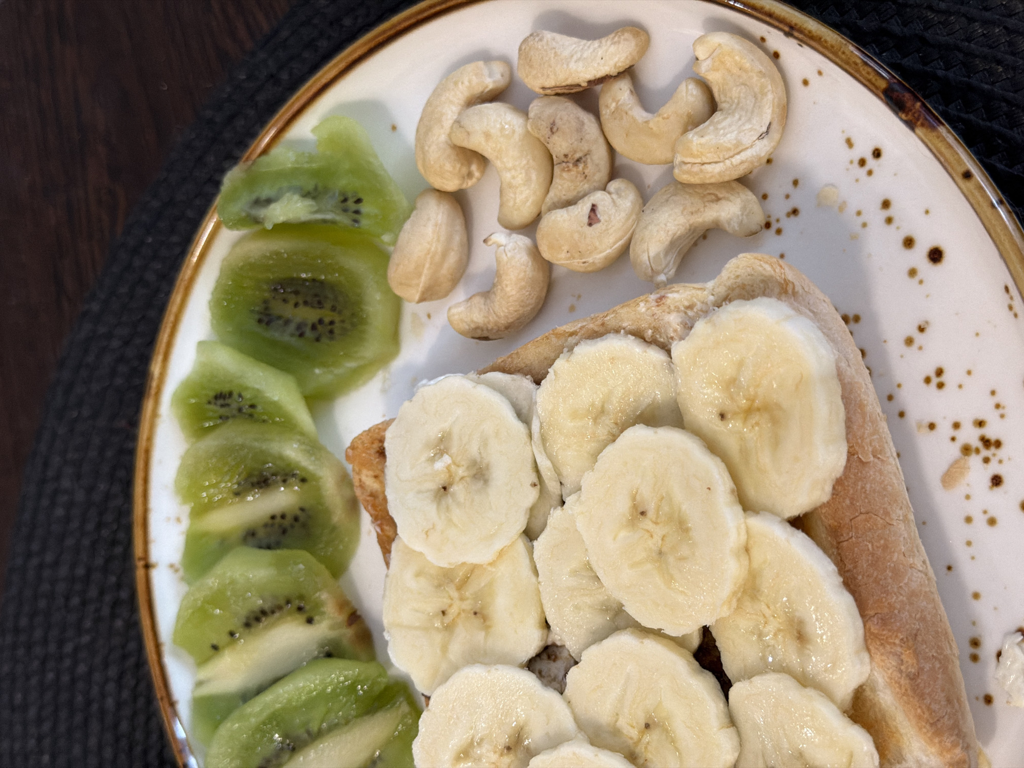 A close-up, high-angle shot captures a healthy meal served on a decorative white plate. The plate, featuring a rustic brown speckled rim and scattered brown speckles on its surface, is placed on a dark, textured mat, which in turn rests on a dark wooden table.
The meal consists of three main components:
1. **Sliced Kiwi:** Arranged along the left side of the plate are multiple thin slices of bright green kiwi, with their characteristic black seeds visible. Some slices show a slight translucency, suggesting they are fresh or perhaps slightly softened.
2. **Cashews:** A small pile of whole, light beige cashew nuts occupies the top central portion of the plate.
3. **Sliced Bananas on Bread:** The lower right of the plate features numerous round slices of light yellow banana, which are laid over what appears to be a piece of toast or bread, mostly covered by the fruit. The bread itself has a light brown, slightly toasted appearance.
The lighting is bright, illuminating the food and highlighting its textures and colors. The image presents a still-life composition of a breakfast or snack, emphasizing fresh fruit and nuts. No people or visible text are present in the image.