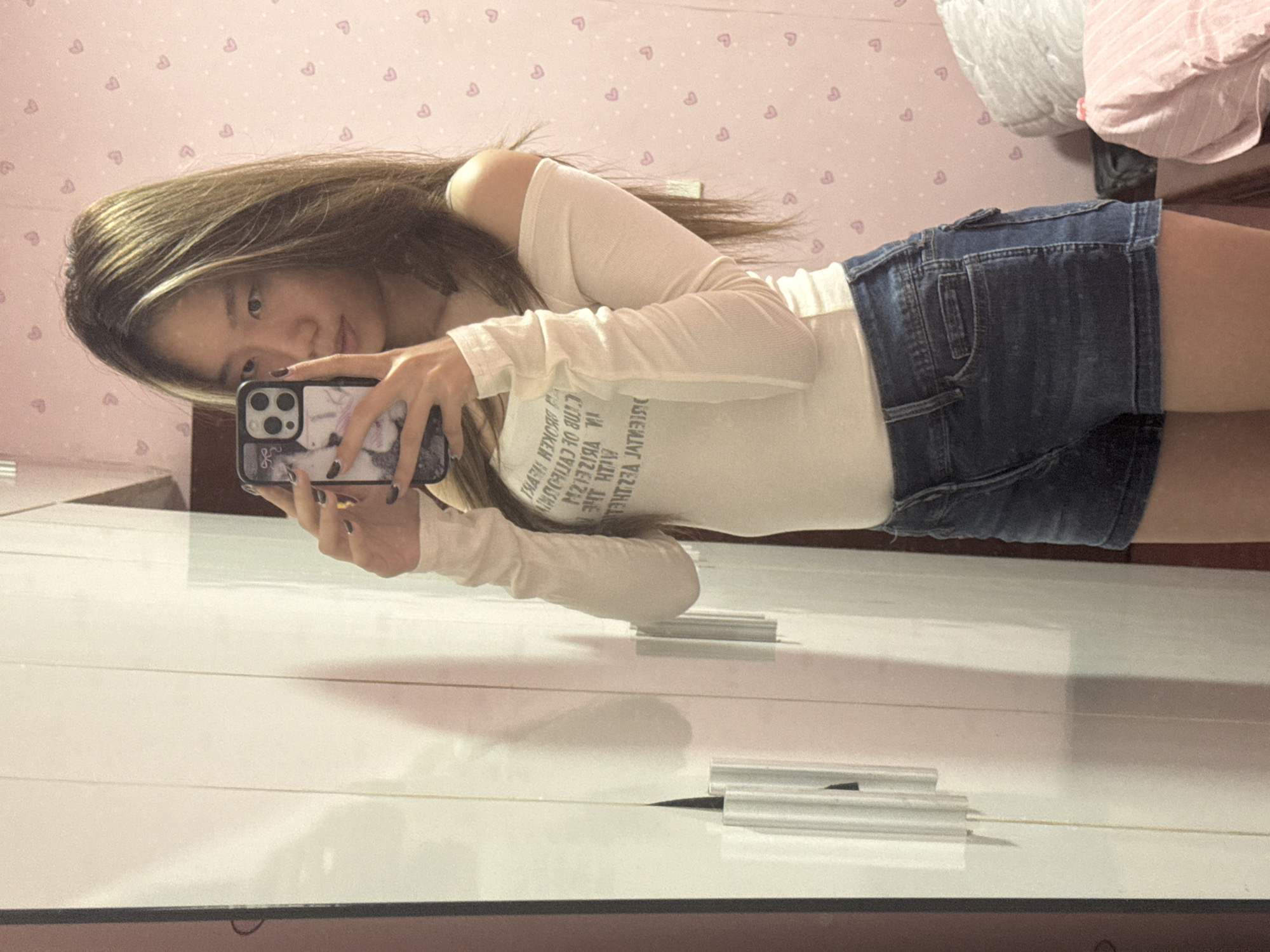 The image shows a young woman in what appears to be a bedroom. She is holding a smartphone and taking a selfie in front of a mirror. She's wearing a white, long-sleeved, off-the-shoulder top and denim shorts. The background includes a pink wall with a heart pattern, and pillows are visible in the upper right corner. The reflection in the mirror shows a white wardrobe or cabinet. The room appears to be in a domestic setting. There is no indication of the location being in Shapingba District, China, or any city.