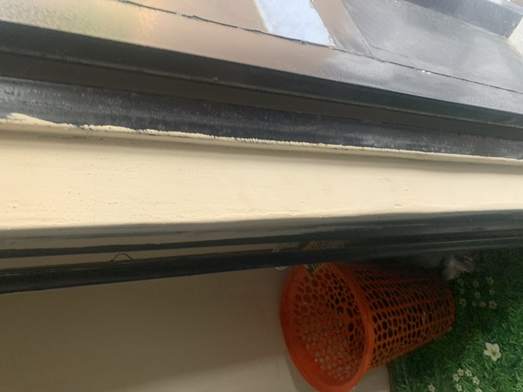 The image shows an orange plastic basket with a mesh pattern on a green, artificial grass mat. The basket is positioned near a black door frame. Part of a wall painted cream is visible to the left of the door frame. The lighting suggests an indoor setting, possibly a doorway or entryway, in Jauro Yinu, Nigeria. No people are visible in the image.
