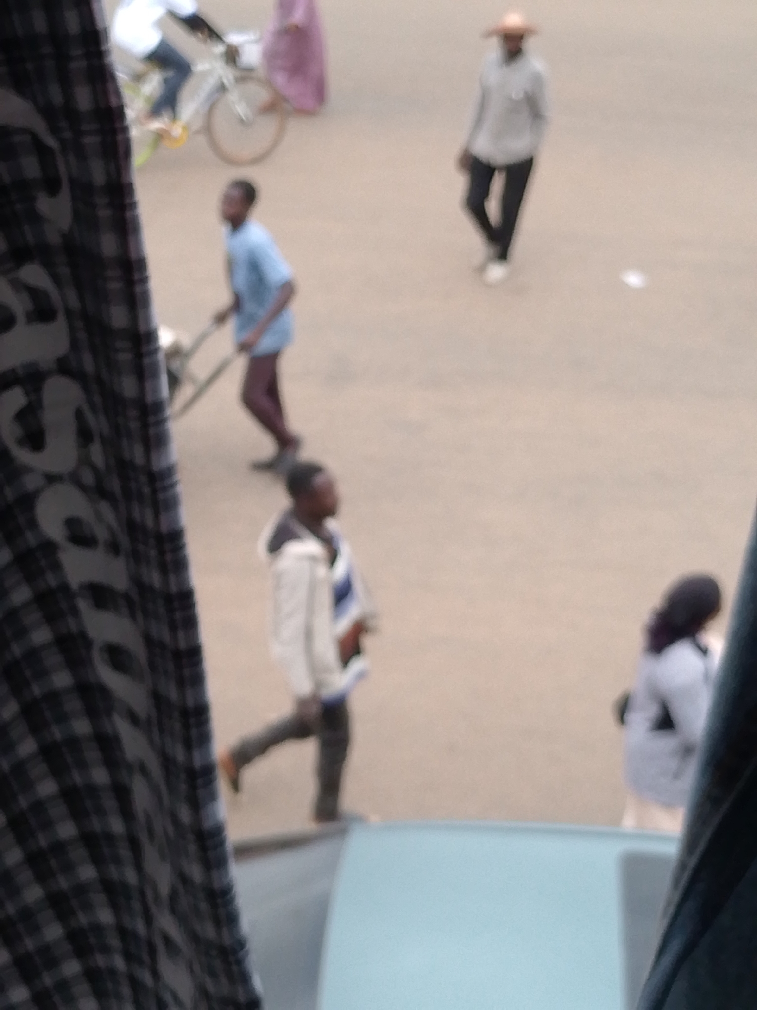 The image, likely taken in Bukuru, Nigeria, shows a street scene viewed from an elevated position, possibly through a bus or car window. The foreground is partially obscured by a plaid curtain. Several people are visible: one person is pushing a handcart, one is walking wearing a hat, another is walking in a white jacket, and another is walking away from the camera wearing a light-colored top. A bicycle is also visible. The ground appears to be a dusty beige color.