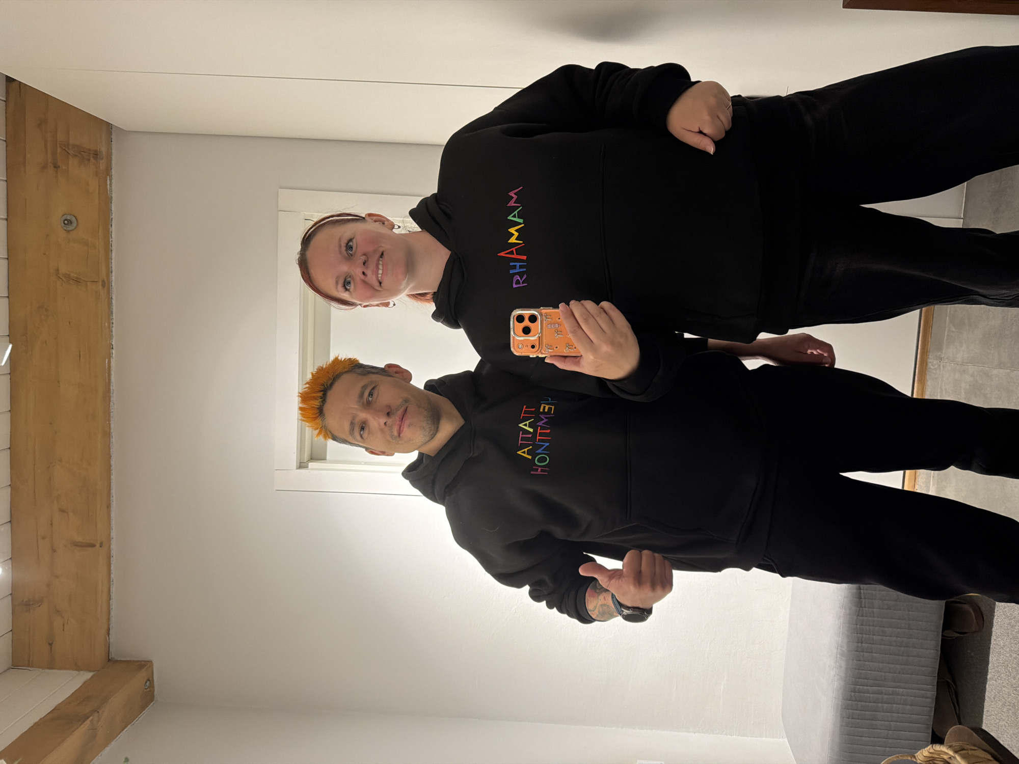 This is a full-body mirror selfie featuring two individuals, a woman and a man, standing side-by-side in an indoor setting in Chaikovsky, Russia. Both appear to be adults and are wearing matching black hoodies with rainbow-colored text.
The woman, positioned on the left, has reddish-brown hair pulled back from her face and is smiling towards the camera. She is holding an orange smartphone with a patterned case in her left hand. The text on her hoodie clearly reads "RHAMAM" in vibrant, multi-colored letters.
The man, on the right, has striking spiky, orange-dyed hair and light facial stubble. He is giving a thumbs-up with his left hand, revealing tattoos on his forearm and a dark-faced watch on his wrist. Due to the mirror reflection, the text on his hoodie appears reversed, reading "ATTATT HONTMEY?" in rainbow colors. He also has a slight smile.
The background consists of plain, light-colored walls. A prominent wooden beam, possibly structural, runs across the upper left and lower left of the frame, adding a rustic architectural element. At the bottom right, a light-colored, possibly upholstered, bench or bed is partially visible. The lighting is bright and even throughout the scene, suggesting an indoor environment with artificial light. Both individuals appear to be in good spirits, posing cheerfully for the photograph.