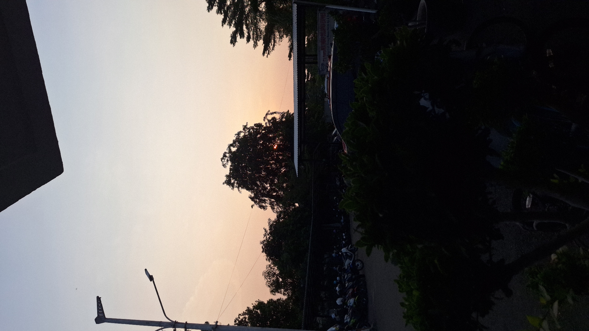 The image depicts an outdoor scene at dusk in Karawang, Indonesia. The sky is a gradient of light blue to orange near the horizon, where the sun is partially obscured by a tree. Silhouetted against the sky are trees, utility poles, and the corner of a building. Below the tree line, a parking area is visible with several motorcycles parked closely together. Vegetation borders the parking area on the right.