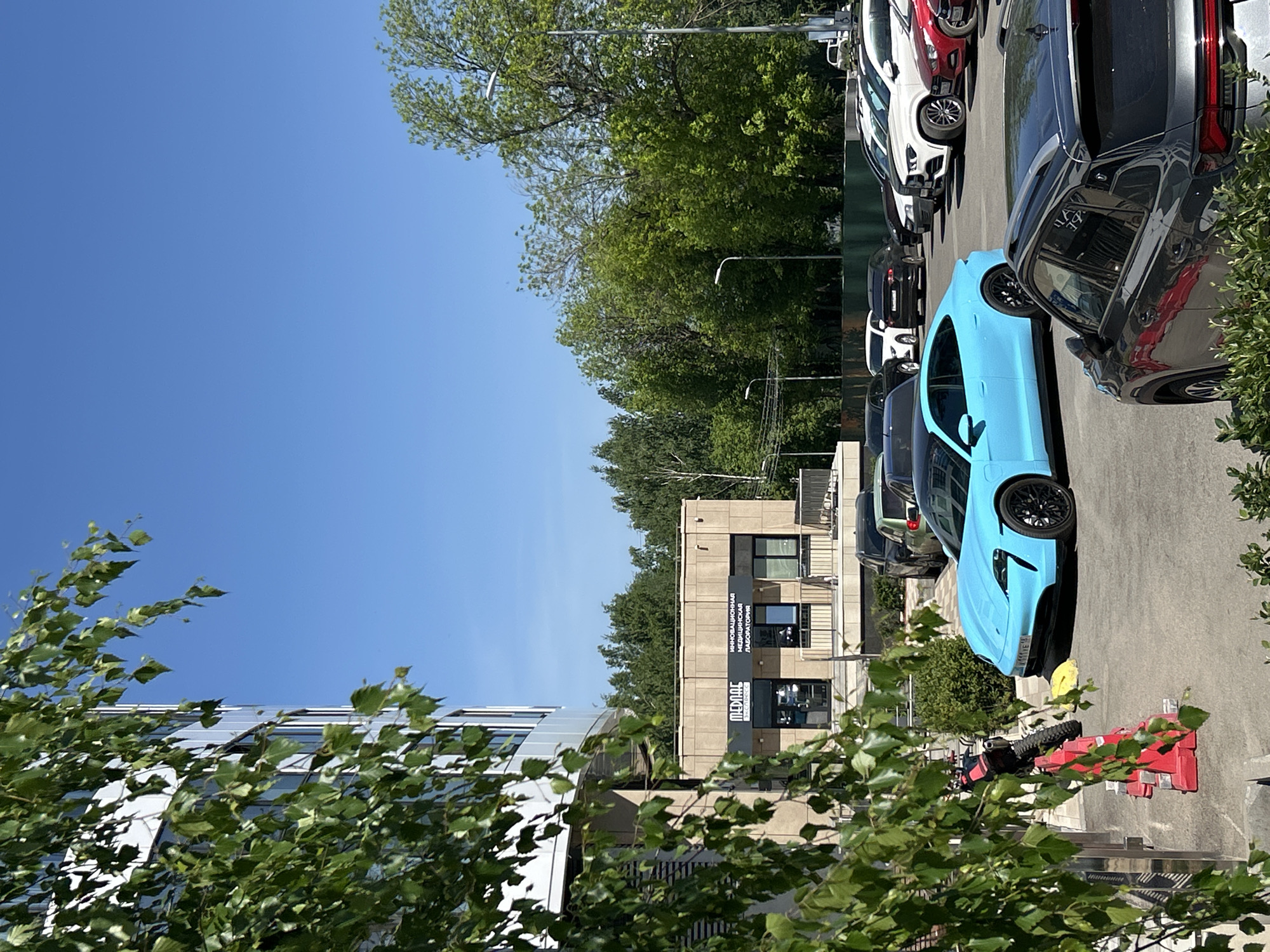 The image shows a parking lot in Izhevsk, Russia, with several cars parked in a row. A bright blue car is a prominent feature in the foreground. Behind the cars, there is a building with a sign in Russian, "МЕДААС" (MEDAAS). The location of the building is near the intersection of st. Krasnogeroiskaya, 104 and st. Lenina, 45, in the Pervomaysky district of Izhevsk. The weather is sunny, with a clear blue sky visible above the trees. A motorcycle is near the blue car, along with red and white traffic barriers. There are trees in the foreground, partially obscuring the view.