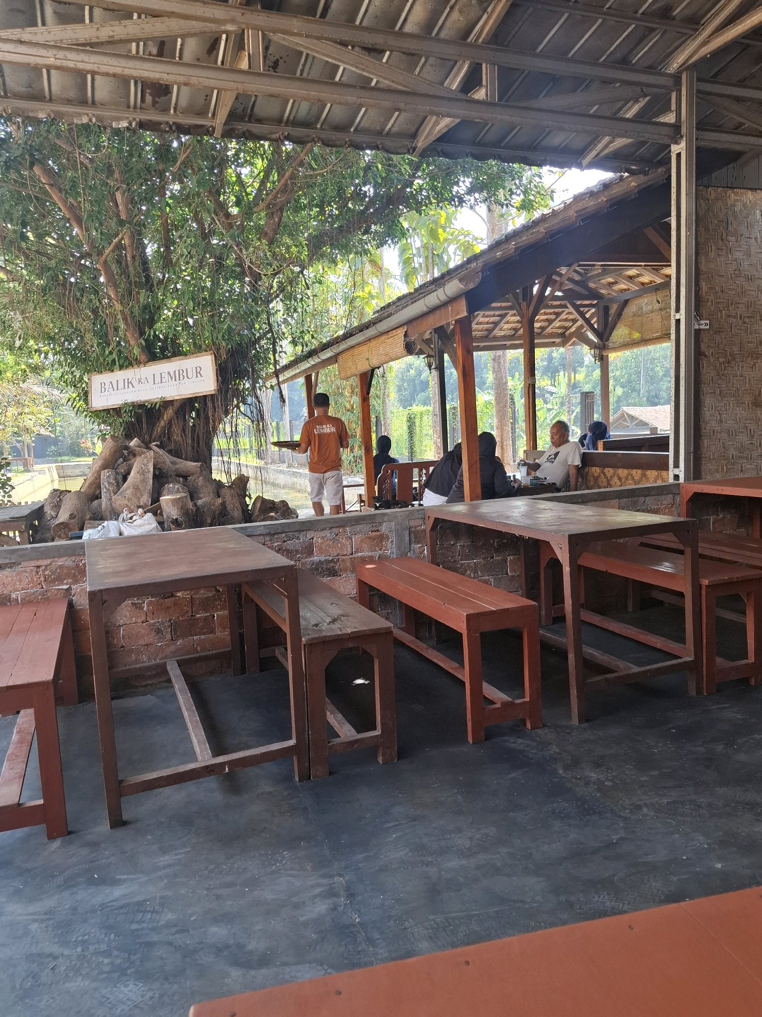 The image depicts an outdoor seating area of a restaurant in Cibentang, Indonesia, with a rustic, natural ambiance. The foreground shows several wooden tables and benches, some stained red, positioned on a dark concrete floor. In the midground, a man in an orange t-shirt with the text "BALIK KA LEMBUR" printed on it is seen holding a tray, walking away from the camera. Behind him, a body of water is visible, bordered by rocks and a large tree with hanging roots. A sign with the same text as the man's shirt is mounted on the tree. In the background, an open-air pavilion with a tiled roof and wooden supports is visible, with several patrons seated at tables. The overall environment suggests a casual dining establishment integrated with nature.
