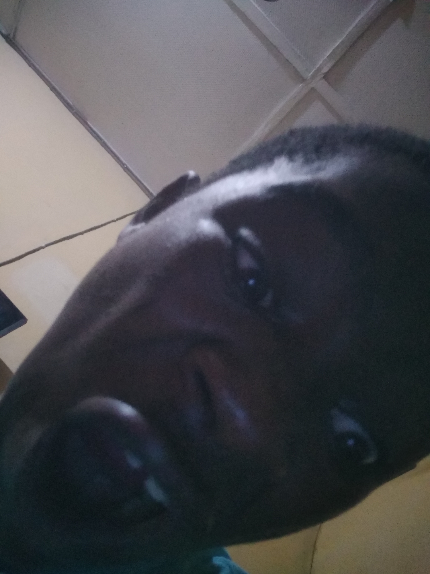 A close-up, angled photograph shows a person's face. The individual has dark skin, short dark hair, and visible eyes and mouth. The lighting is dim, with shadows obscuring some facial features. The background consists of a ceiling with a textured surface and geometric patterns. A portion of what appears to be a wall or other surface is visible in the lower right corner.